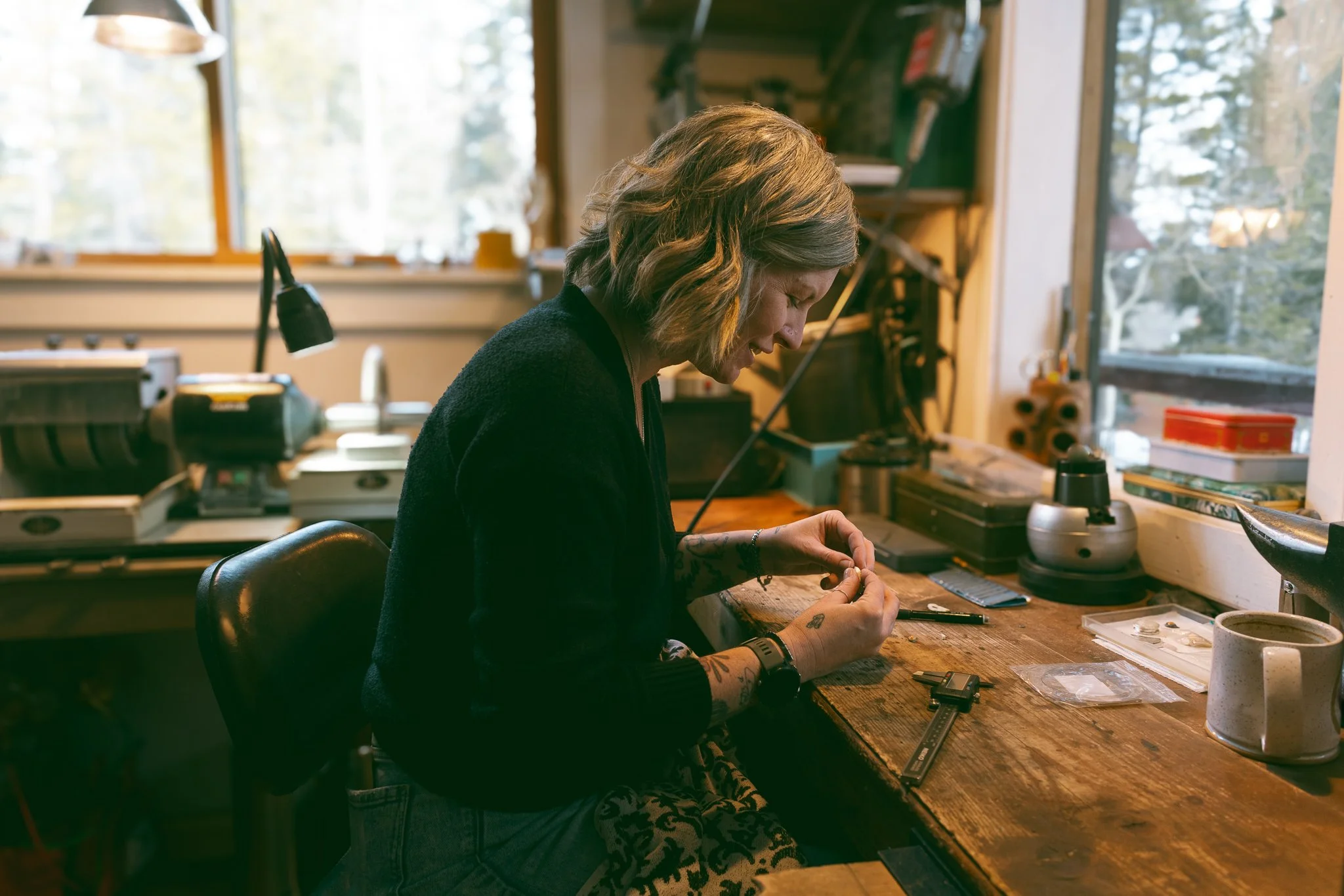 Person working at a wooden workbench in a workshop, inspecting a small object with tools and a cup nearby, natural light coming from a window.
