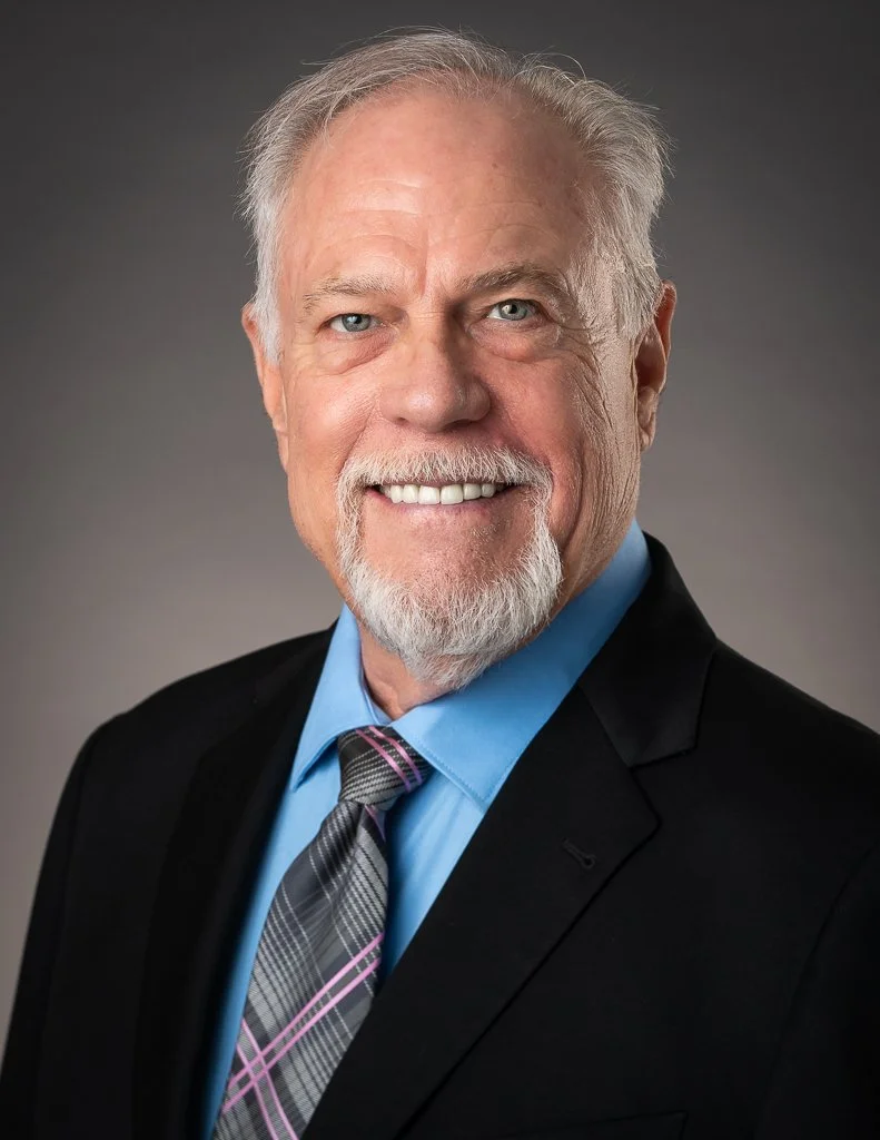 Smiling older Real Estate Broker with white hair, beard, suit, blue shirt, and plaid tie posed against a blue background.