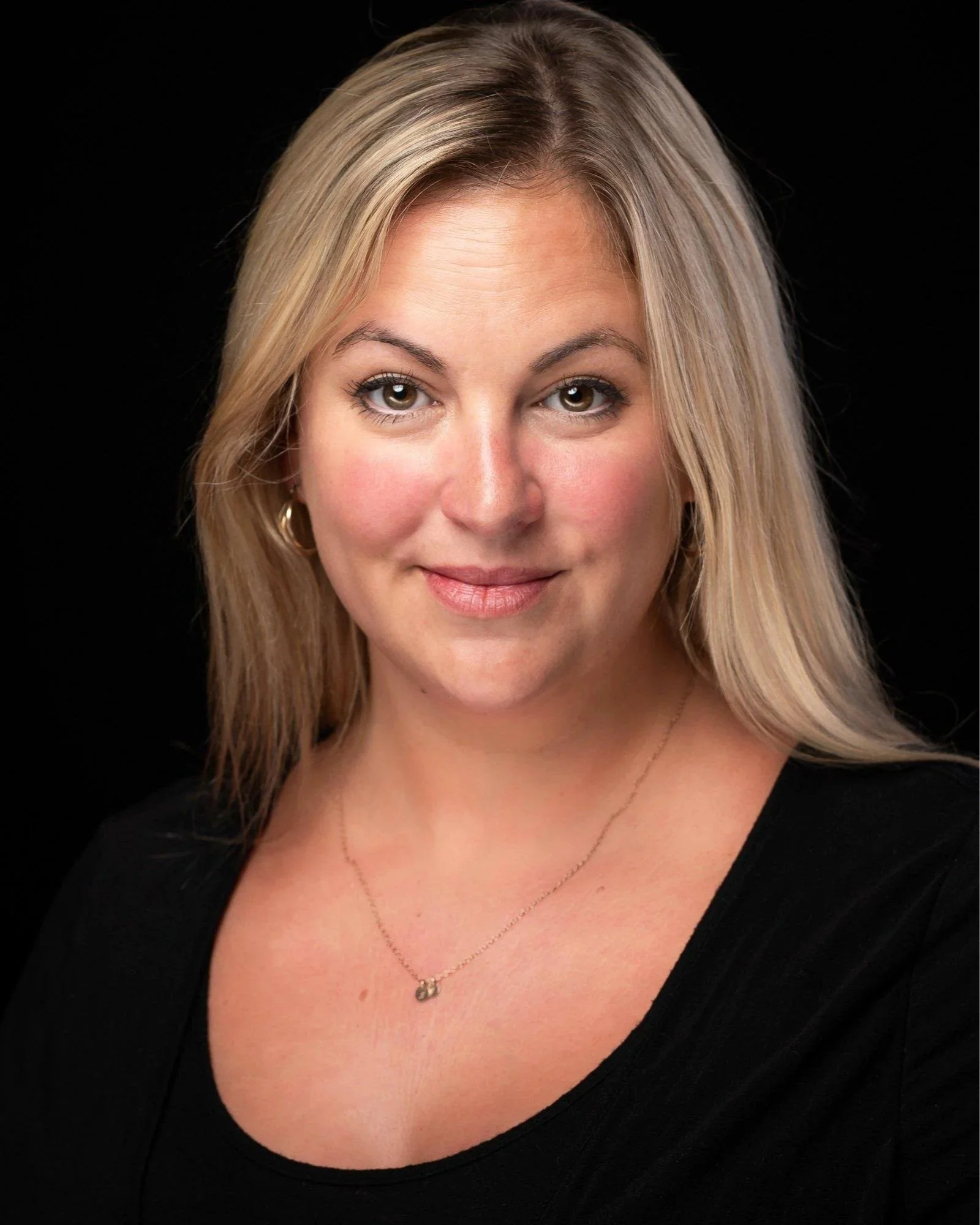 A Team Manager with long blonde hair, in a black top and necklace, poses against a black background, smiling slightly.