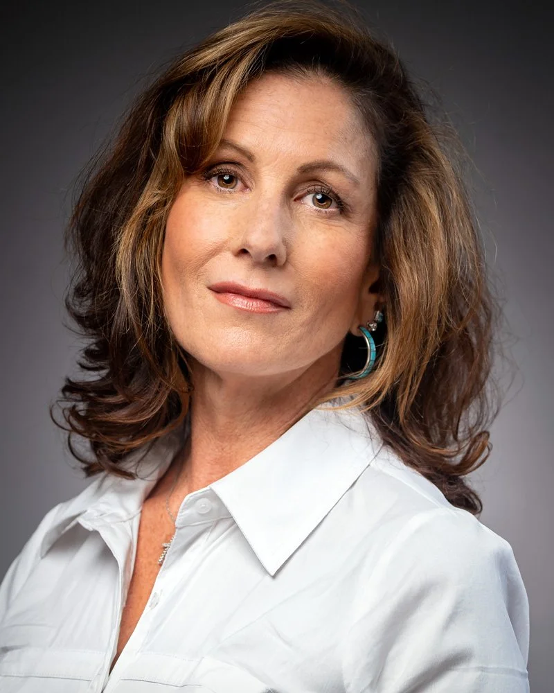 Business owner with wavy brown hair wearing a white blouse, looking at the camera with a neutral expression, gray background.