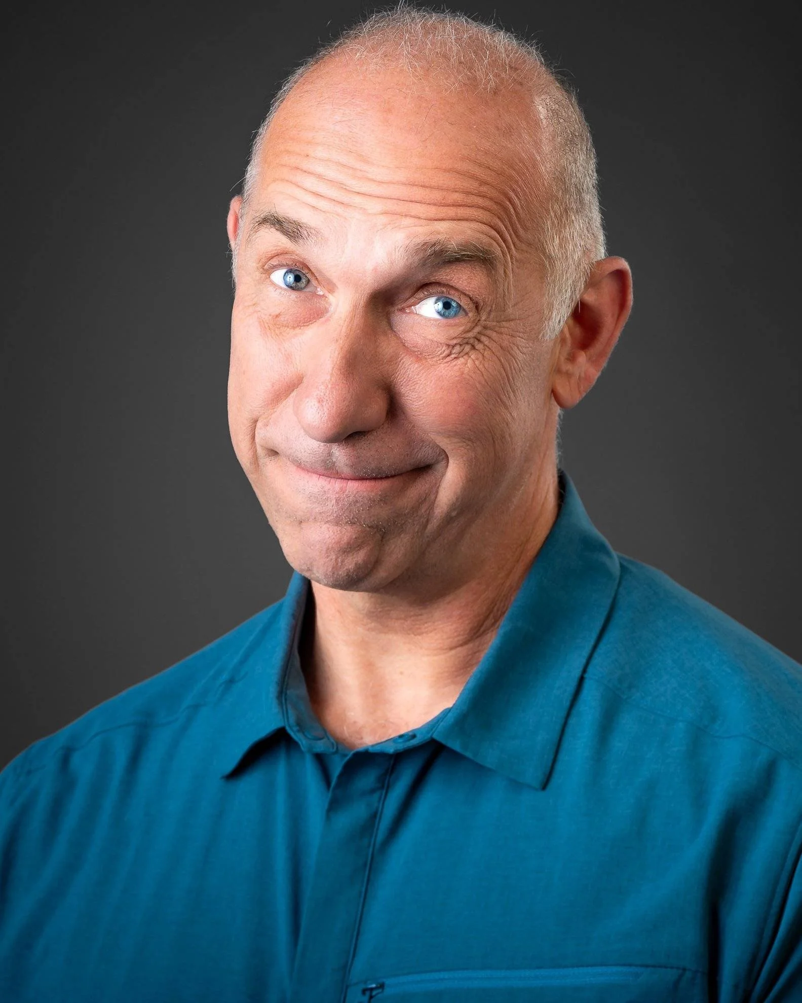 Bald man in a teal shirt flashes salesman humor with a smile and raised eyebrows against a plain dark background.