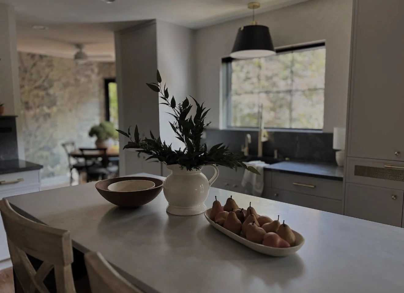 A kitchen with a white countertop, a white pitcher with green leaves, a bowl, and a plate of pears. In the background, windows let in natural light, and there is a dining area with a table and chairs.