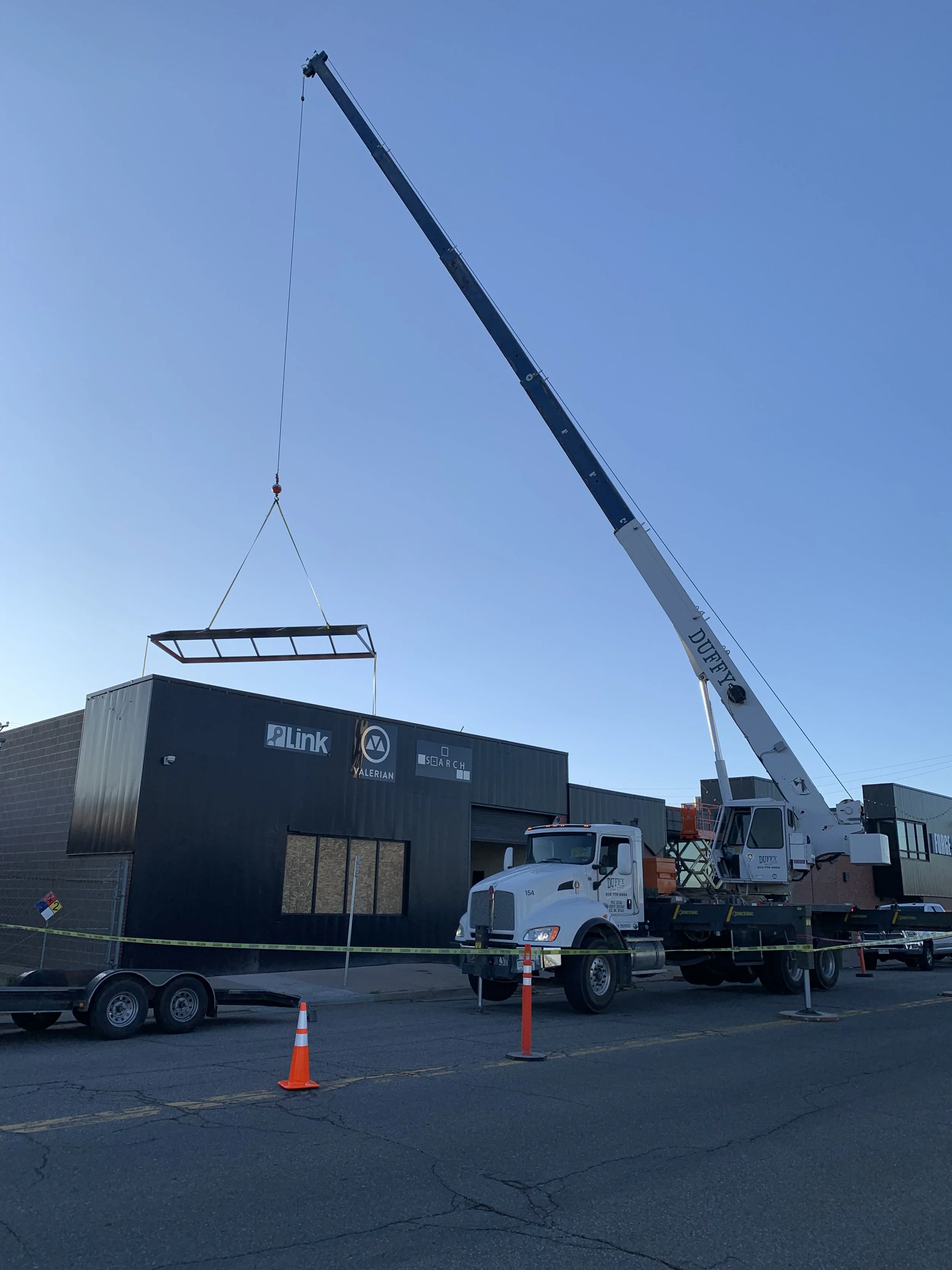 A crane lifting a large metal frame in front of a construction building during daytime with orange traffic cones and a trailer in the foreground.
