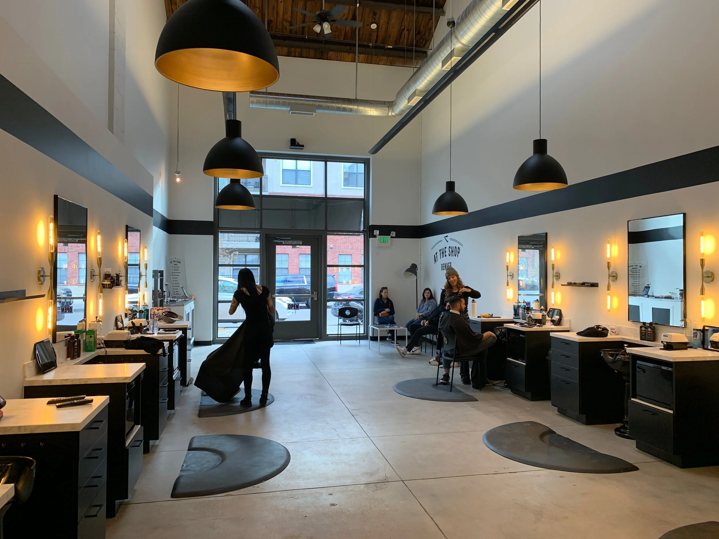 Interior of a modern coffee shop with people sitting and a barista preparing drinks. Black pendant lights hanging from a wooden ceiling, large windows in the background, and a black and white color scheme inside.