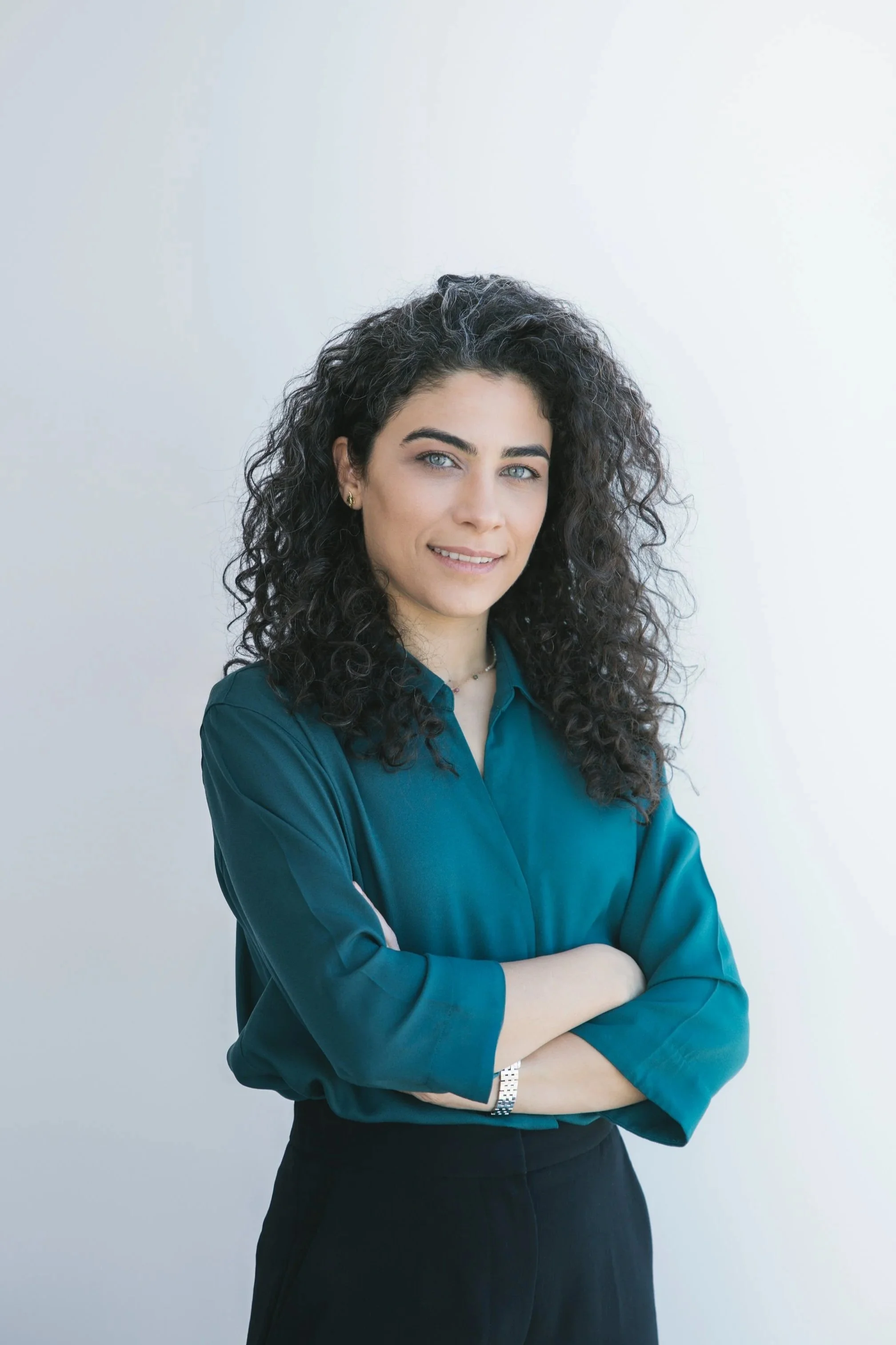 A young woman with curly dark hair, blue eyes, wearing a teal blouse and black pants, standing with arms crossed and smiling against a plain light background.