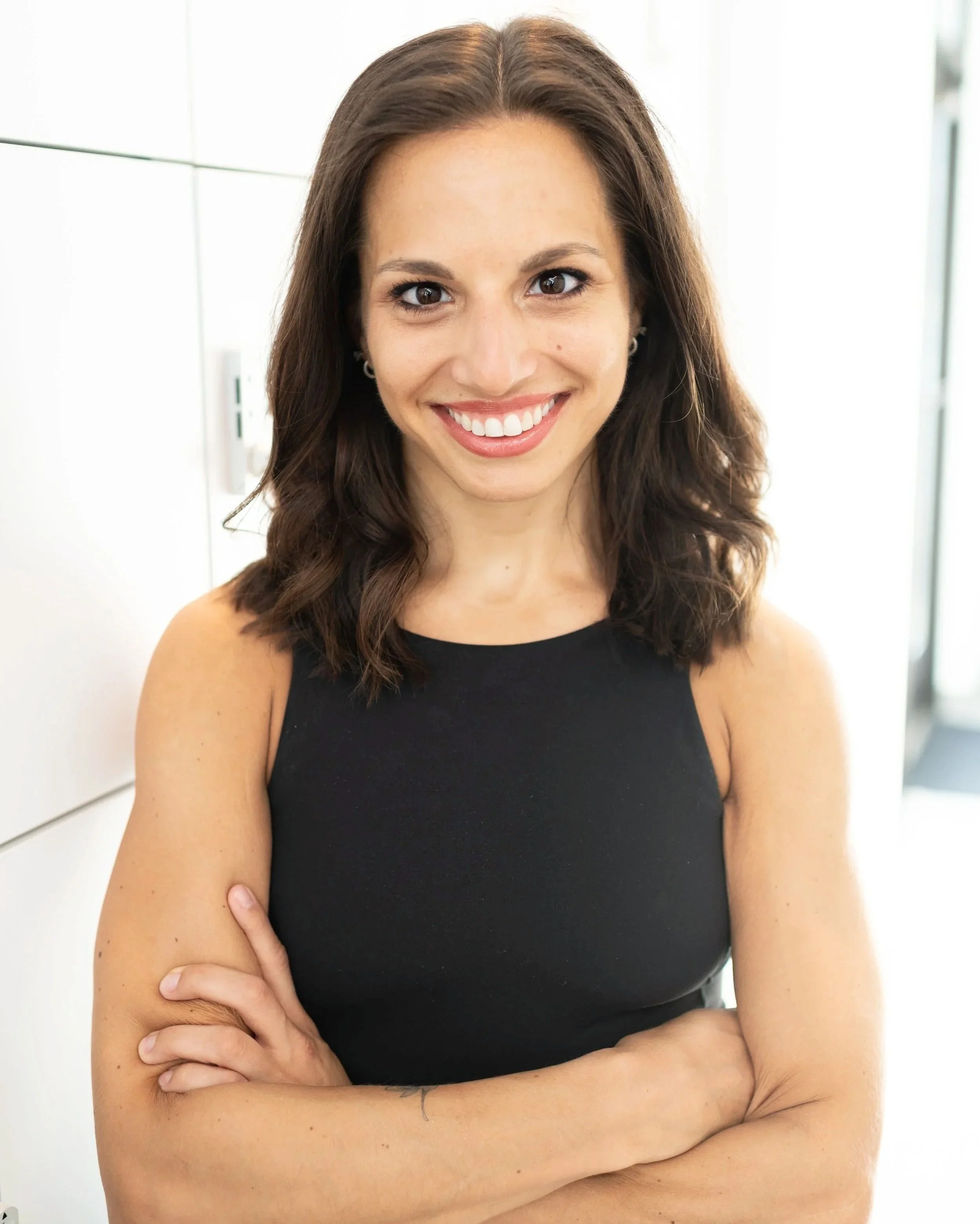 Close-up of a woman with long brown hair, smiling, wearing a black sleeveless top, standing indoors with white walls.