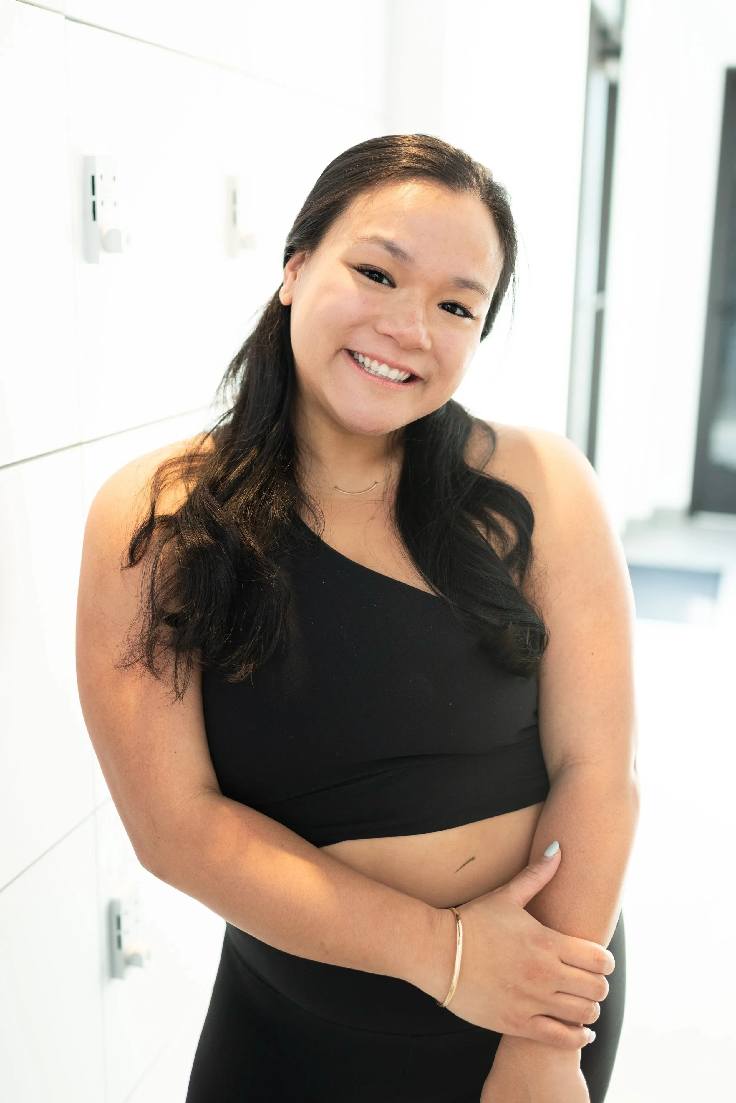 Young woman with long dark hair smiling, wearing a black crop top, standing indoors near white lockers.