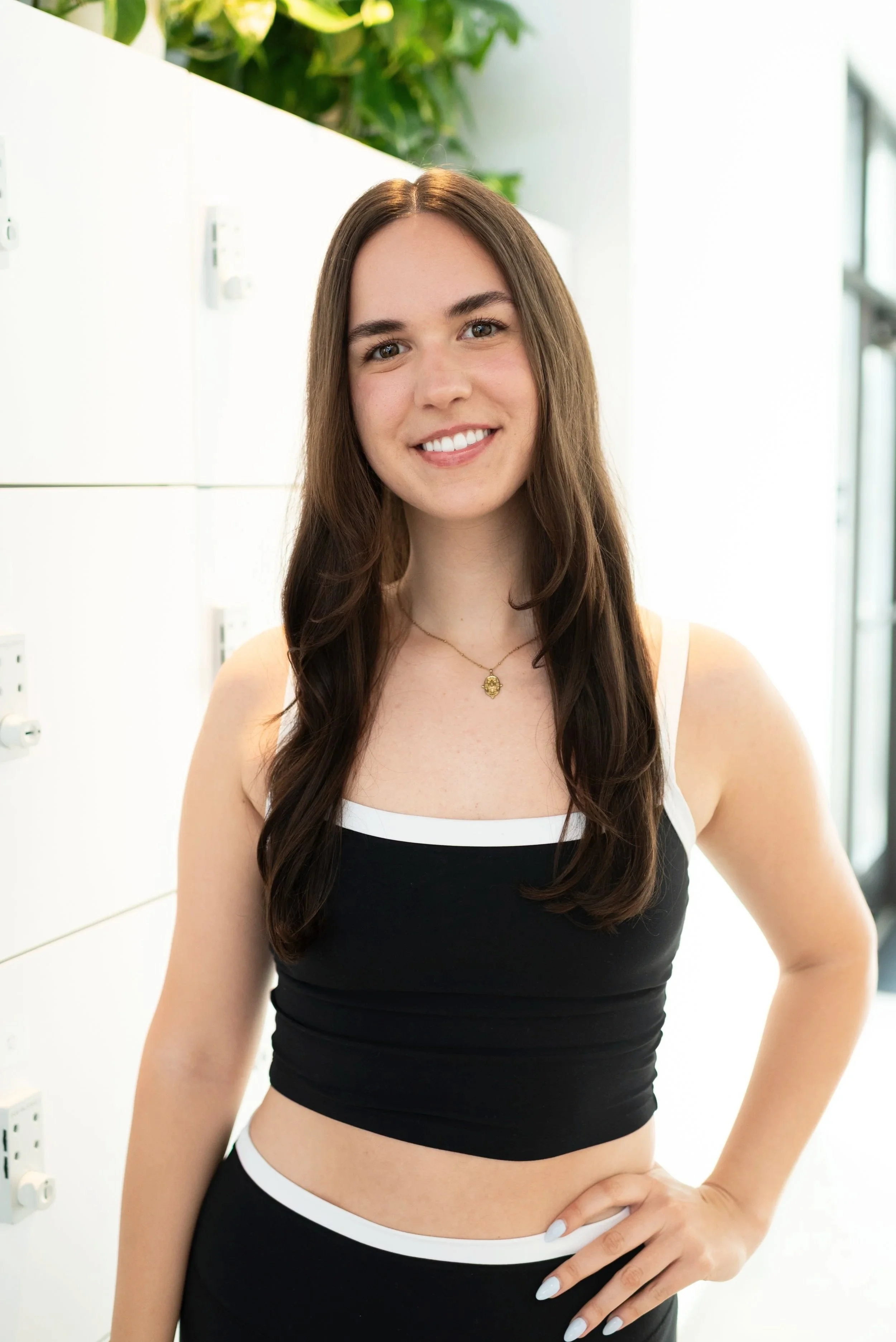 A young woman with long brown hair and light skin smiling, wearing a black and white sleeveless top and a gold necklace, standing indoors near white cabinets and greenery in the background.