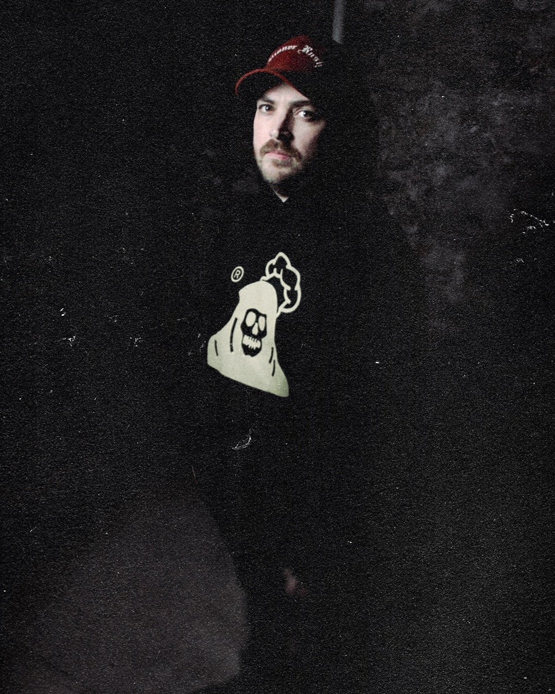 A man with facial hair wearing a black hoodie with a skull graphic and a red and black baseball cap, standing against a dark background.