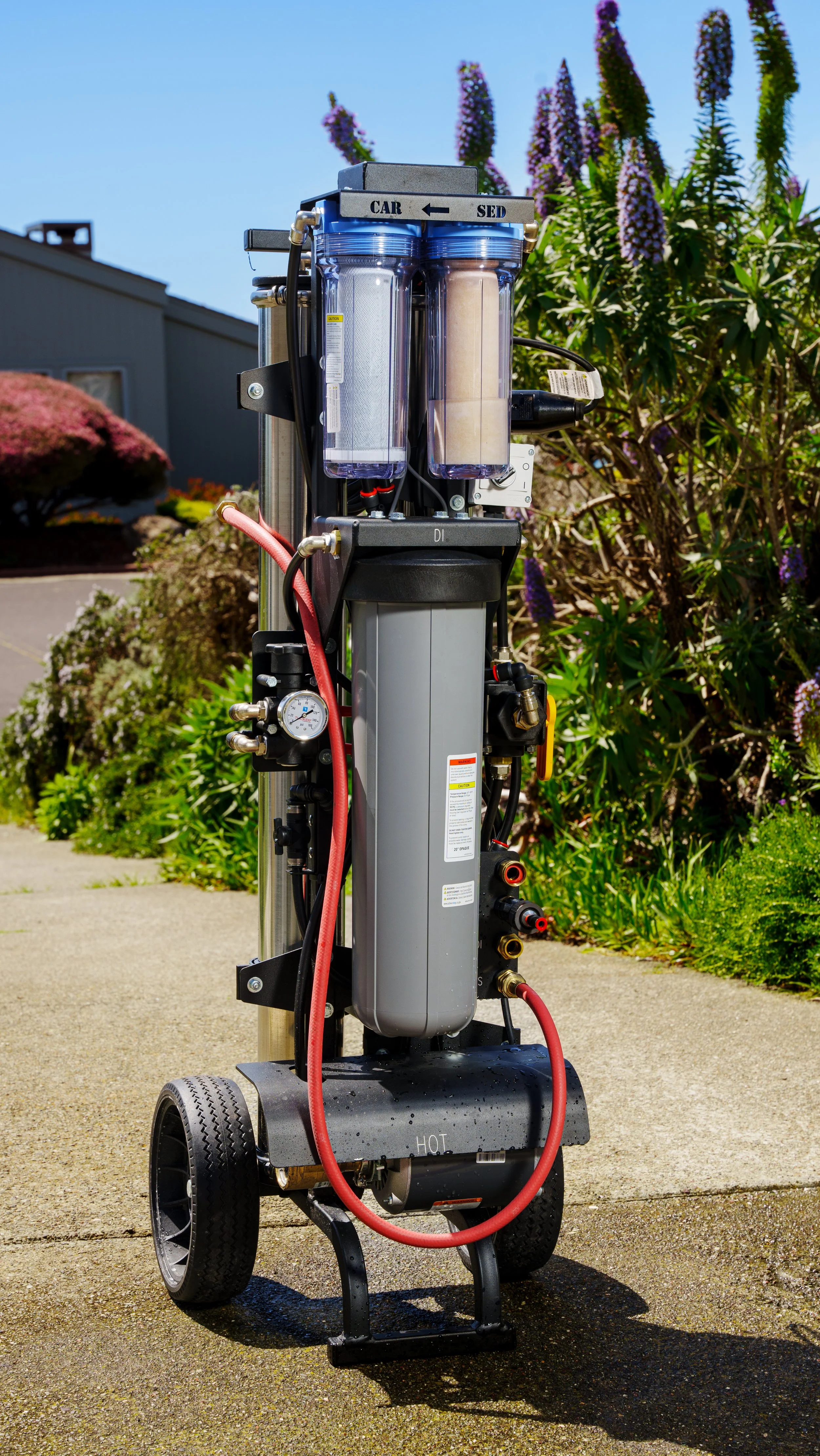 A mobile water filtration system with two clear cylindrical filters, hoses, and gauges, labeled for car and sink use, set outdoors on pavement with plants and a building in the background.