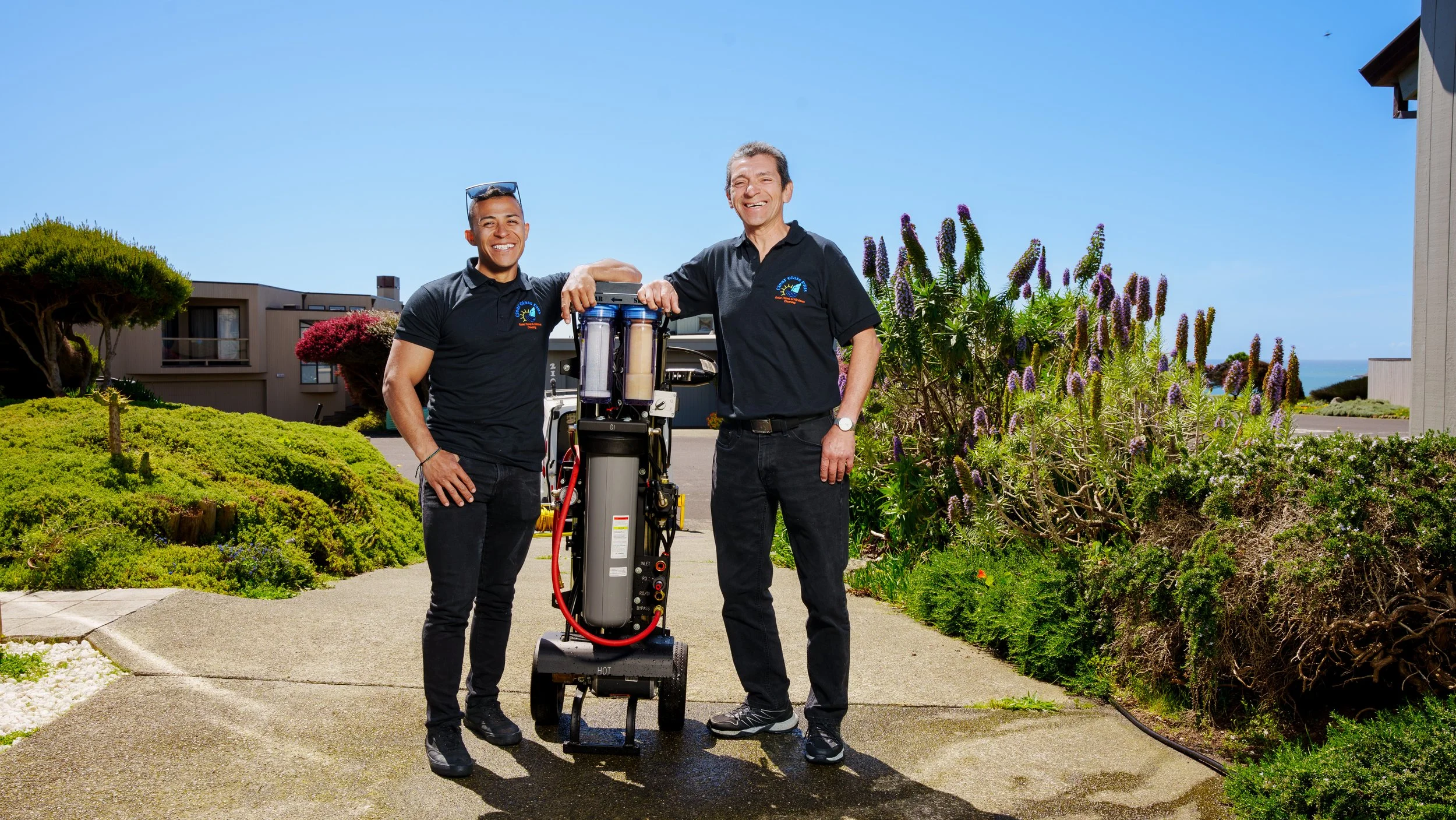 Two smiling men in black uniforms standing outside on a sunny day with a water purification system. They are surrounded by lush green plants and colorful flowers, with residential buildings and a clear blue sky in the background.