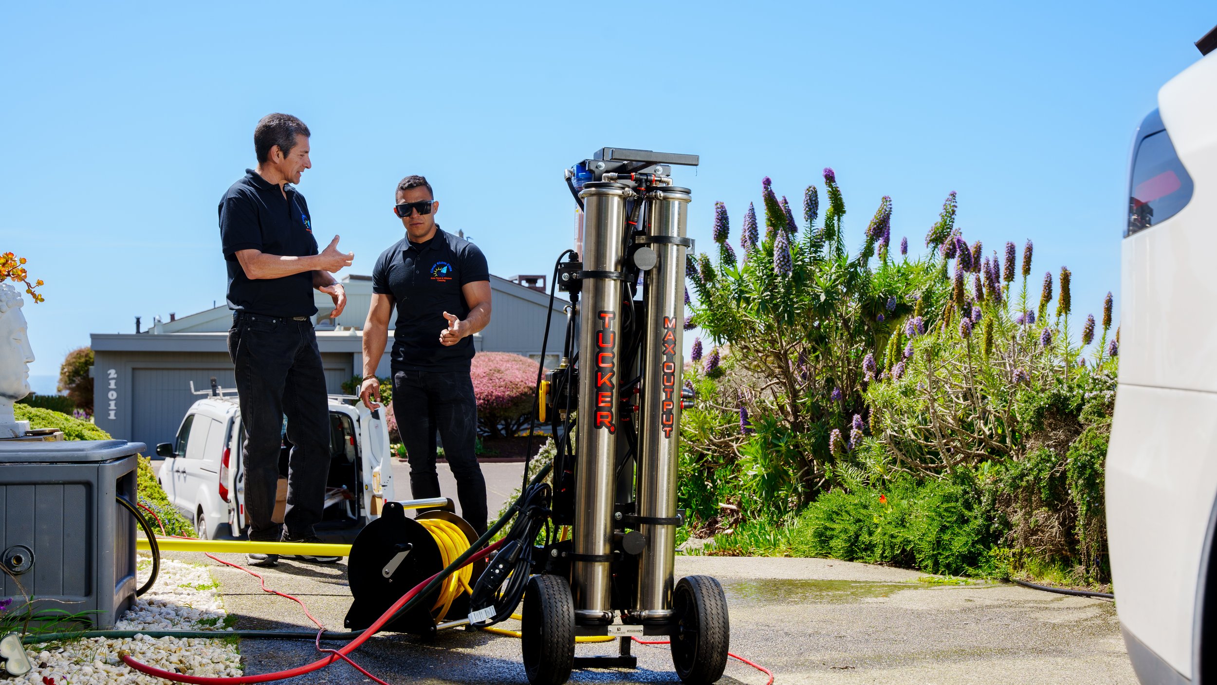 Two men in black shirts are working outdoors with high-tech equipment near flowering plants and a parking lot. One man is explaining while the other listens, with a vehicle and a building in the background.