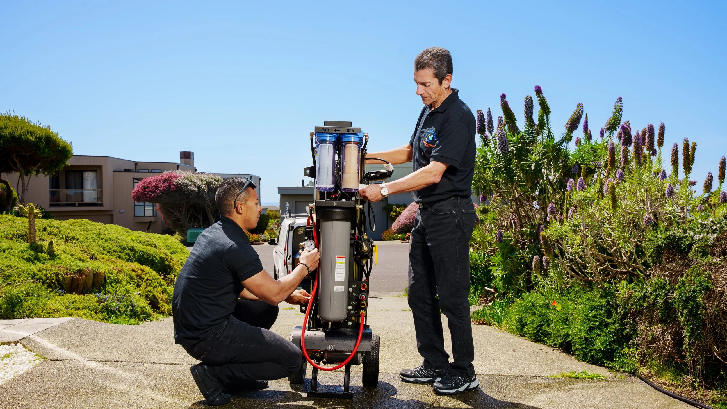 Two technicians working on a piece of equipment outdoors, with vibrant greenery and residential buildings in the background.
