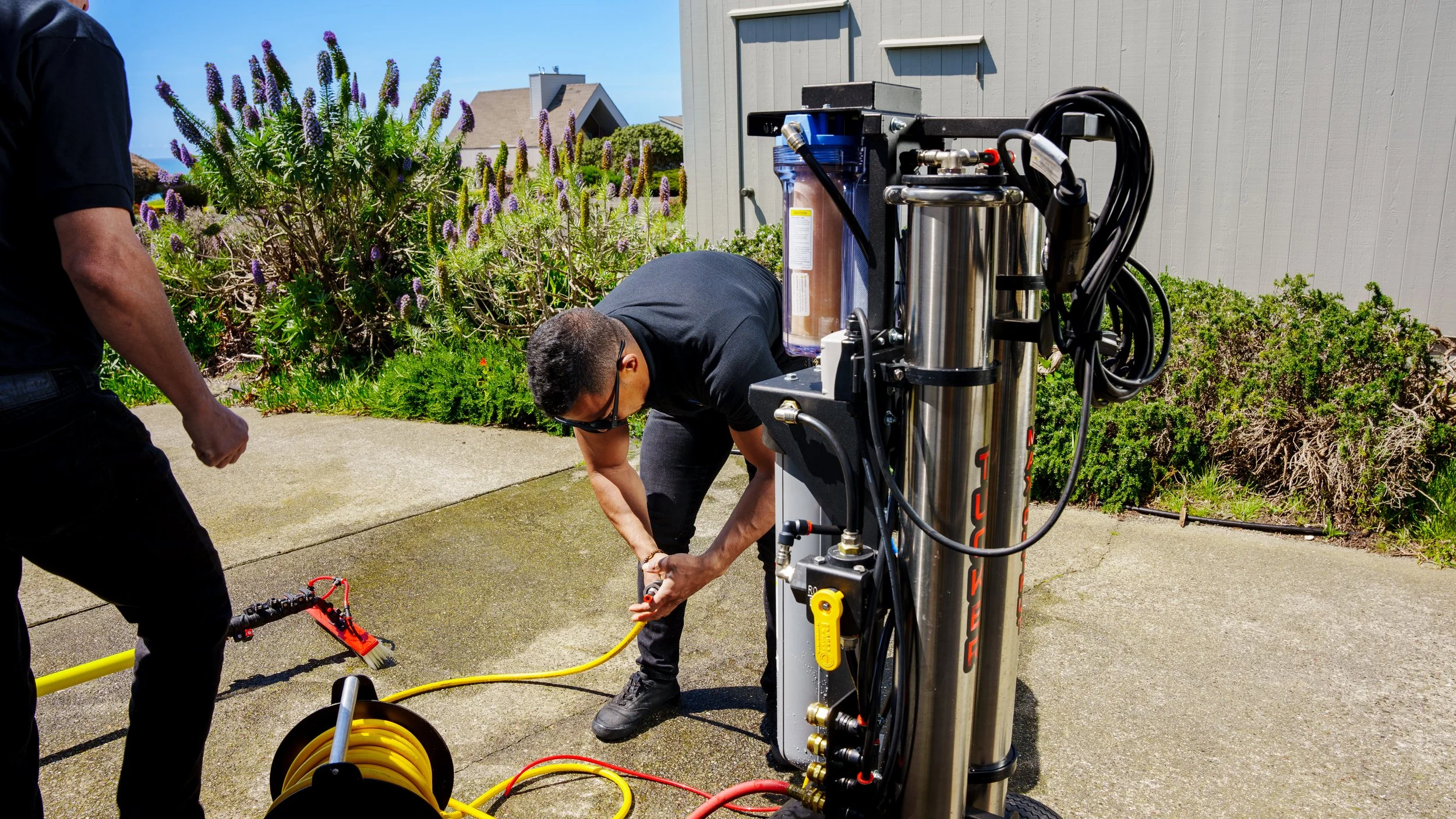 Two technicians installing or repairing a water filtration or reverse osmosis system outdoors on a concrete sidewalk, with plants and houses in the background.