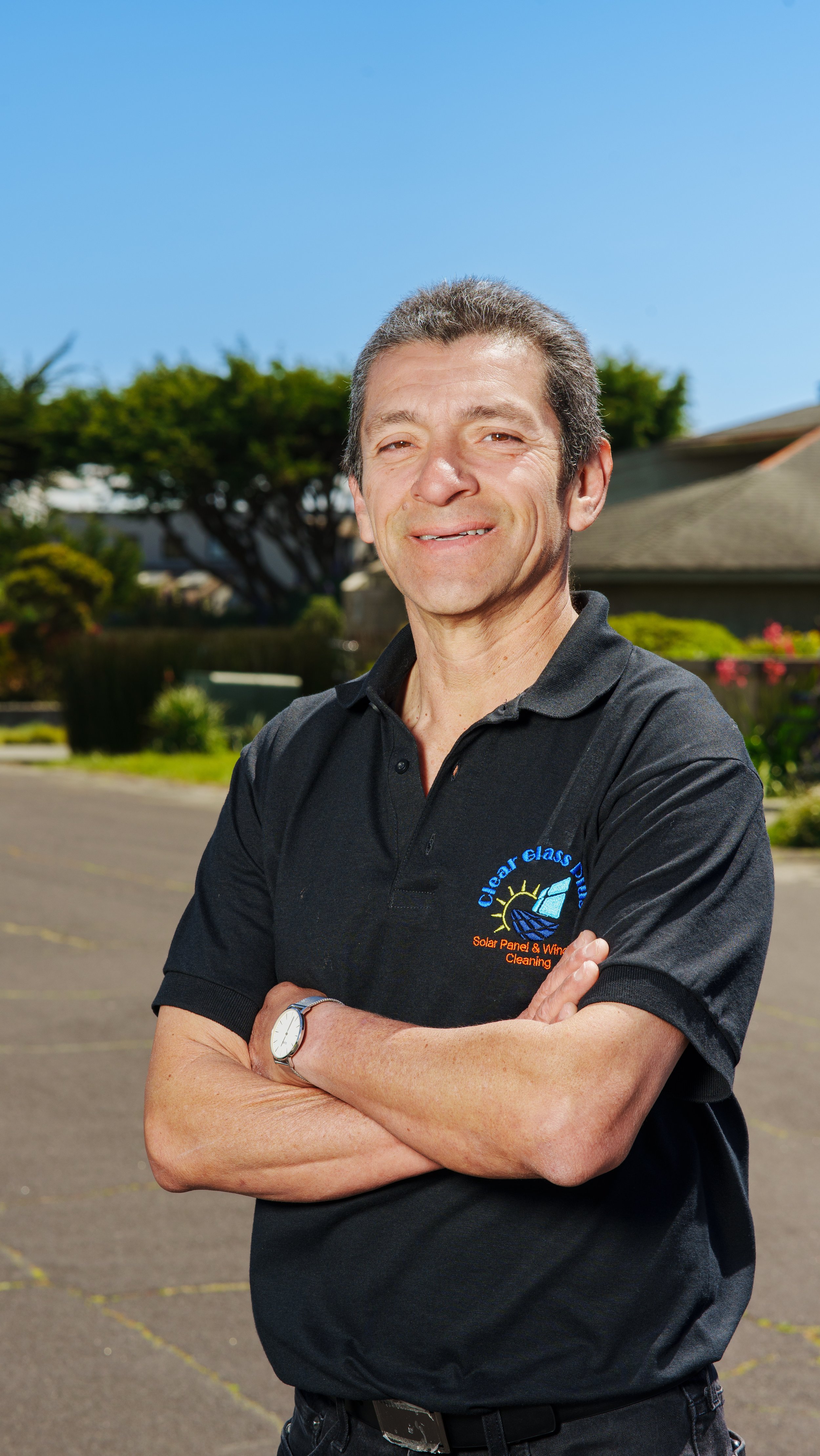 Smiling man with gray hair standing outdoors with crossed arms, wearing a black polo shirt with logo, in front of trees and a building under a blue sky.