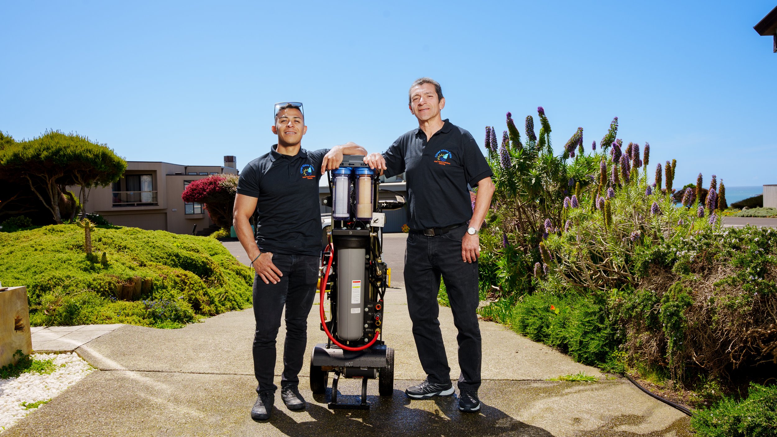 Two men standing outdoors by a water filtration system, with plants and buildings in the background under a clear blue sky.