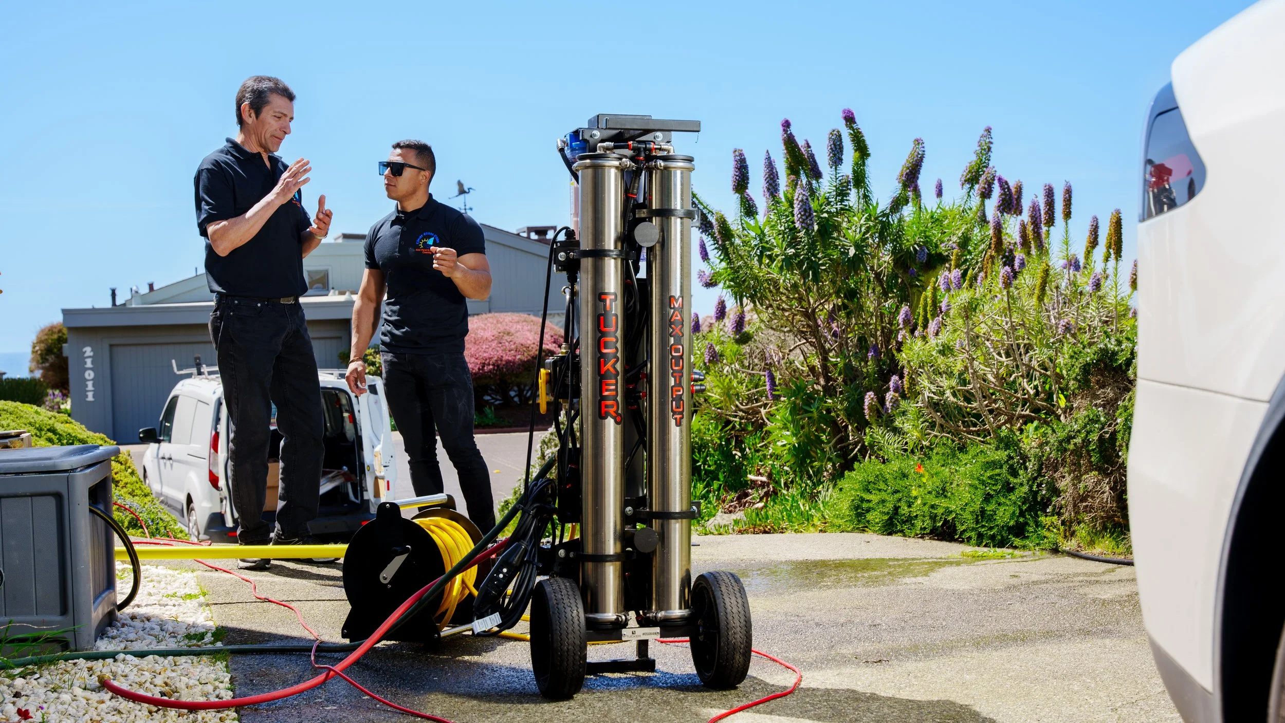 Two men in black shirts working outdoors with a submersible pump labeled "Tucker Maxco" on a sunny day, surrounded by greenery, flowering shrubs, and vehicles.