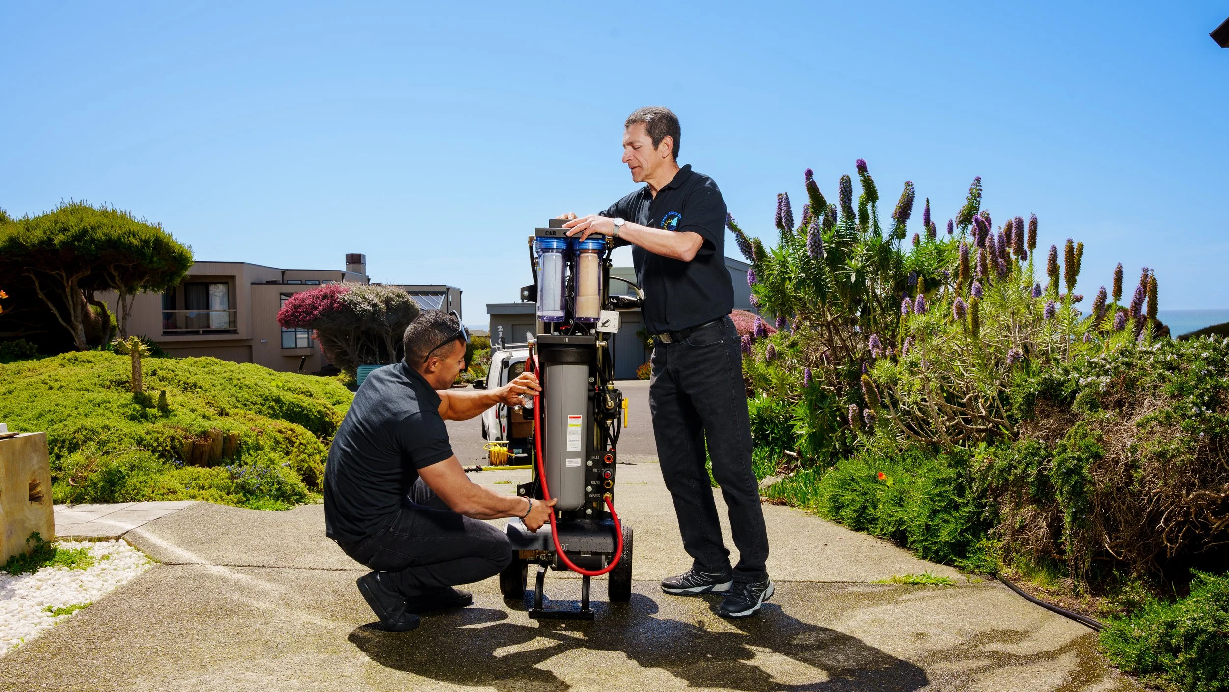 Two men working on a water filtration system outdoors in a landscaped area with flowering plants, trees, and residential buildings in the background.