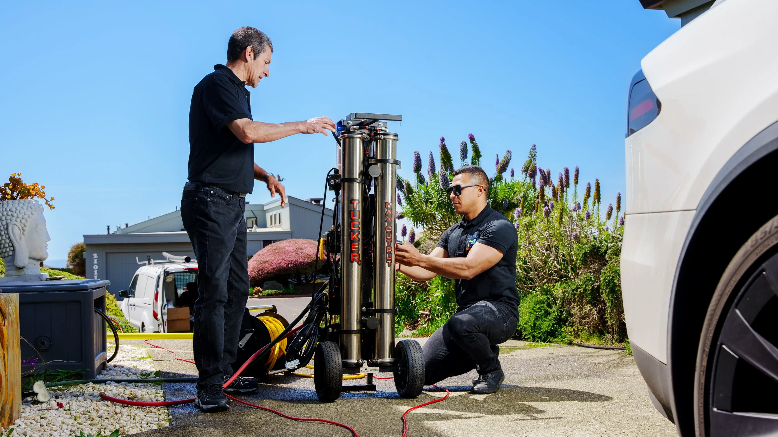 Two technicians working on a large, vertical, metallic equipment labeled 'Kicker Max Output' outdoors near a white vehicle. One is standing and the other is kneeling, both focused on the equipment, with a blue sky and landscaping in the background.