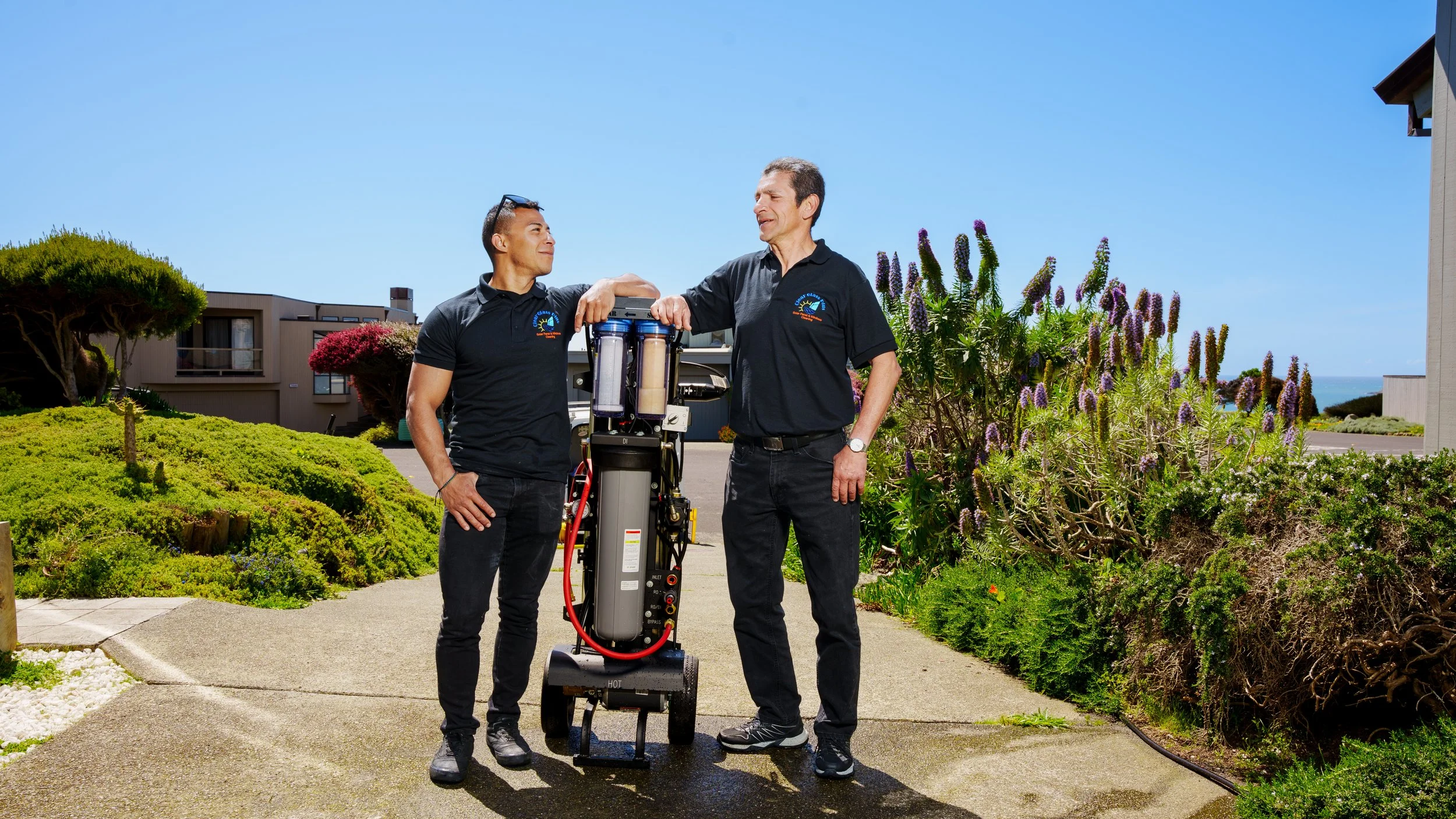 Two men standing outdoors in a garden, having a conversation, with a water filtration or purification system between them, under a clear blue sky.