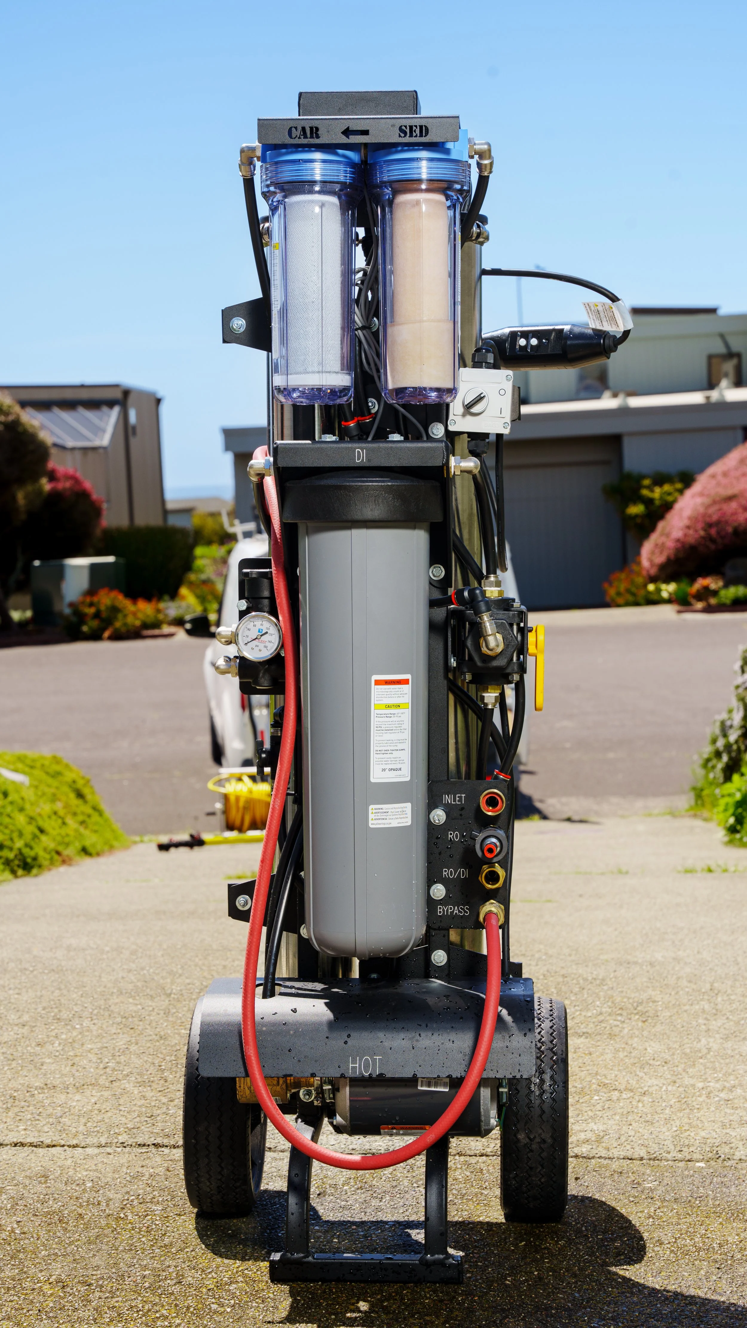 A reverse osmosis water filtration system mounted on a wheeled cart, with two clear filtration canisters, connected pipes, gauges, and a control knob set outdoors on a driveway with houses and bushes in the background.