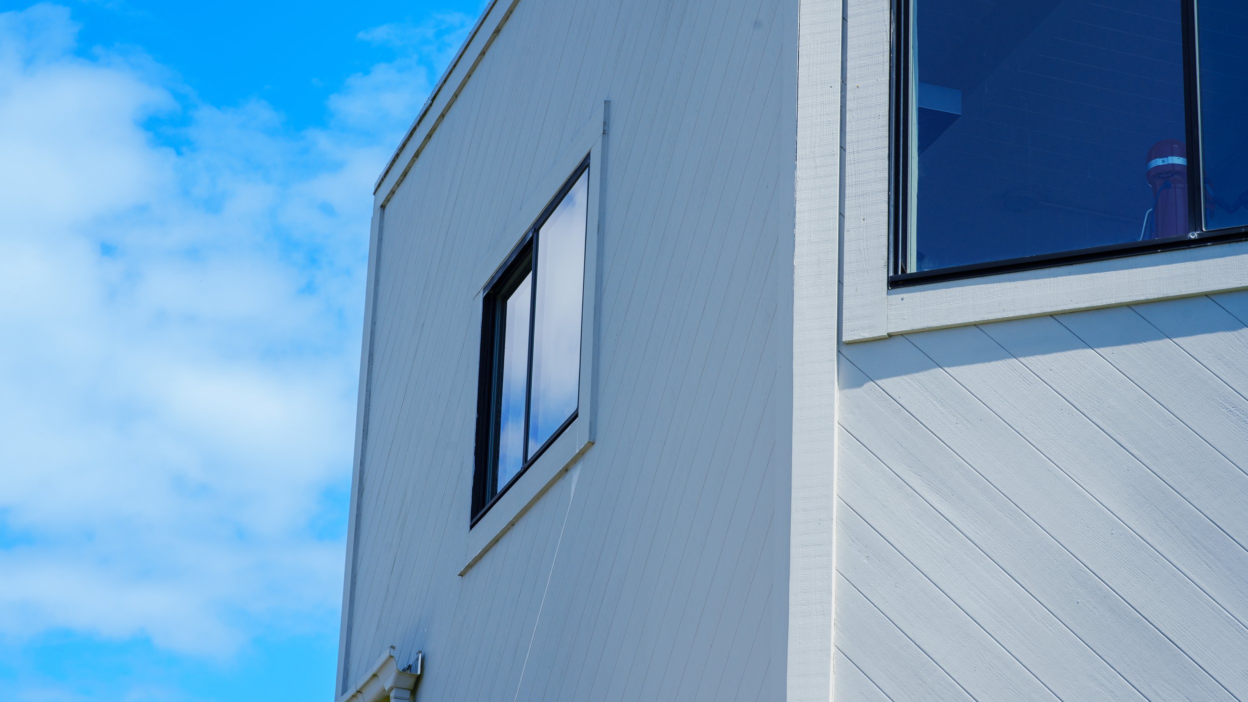 A close-up of a modern building with white horizontal siding and large, dark-framed windows against a bright blue sky with some clouds.