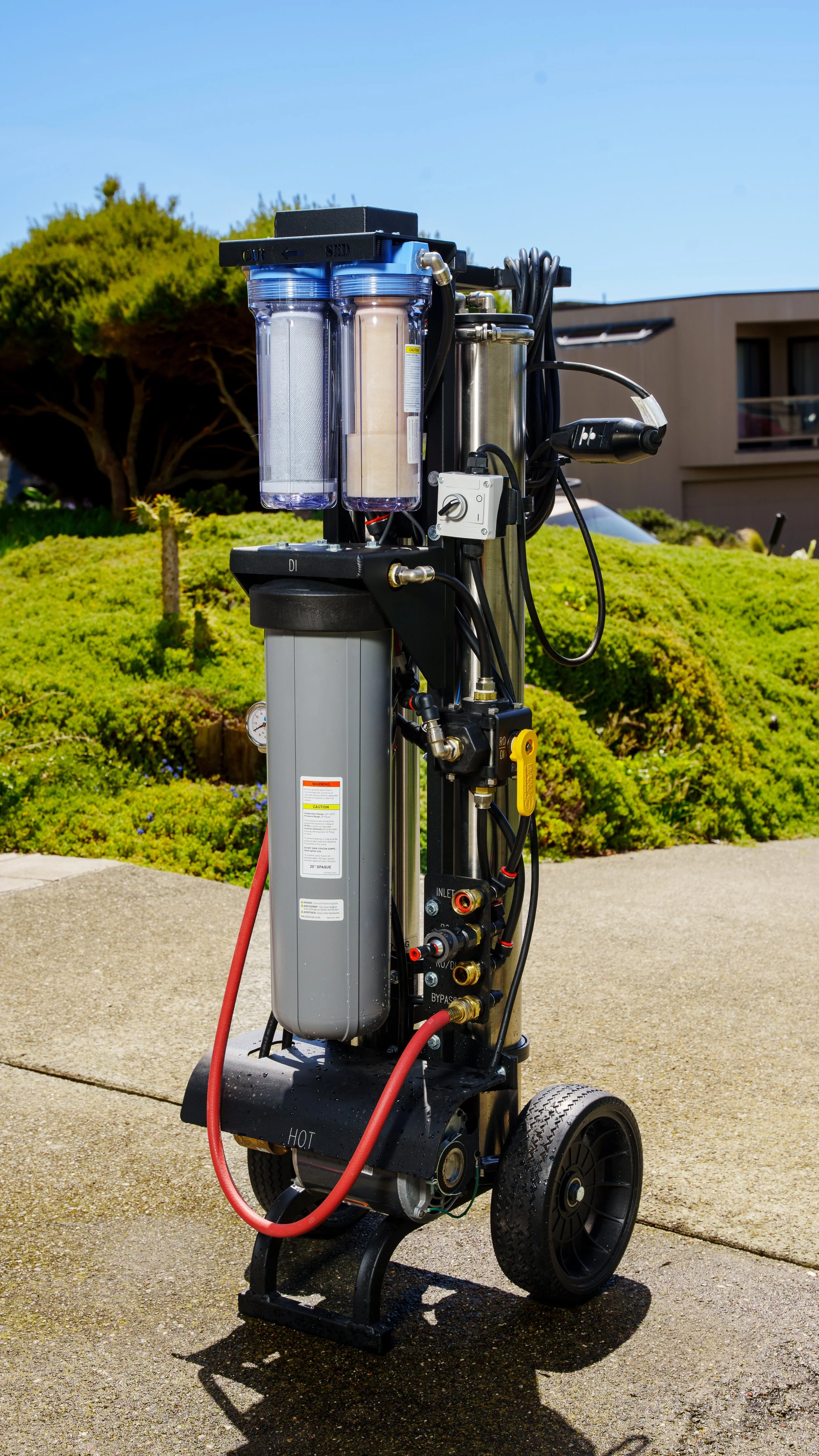 A portable water filtration system with two clear filter containers, connected hoses, and a control panel, placed outdoors on a sidewalk with greenery and a building in the background.