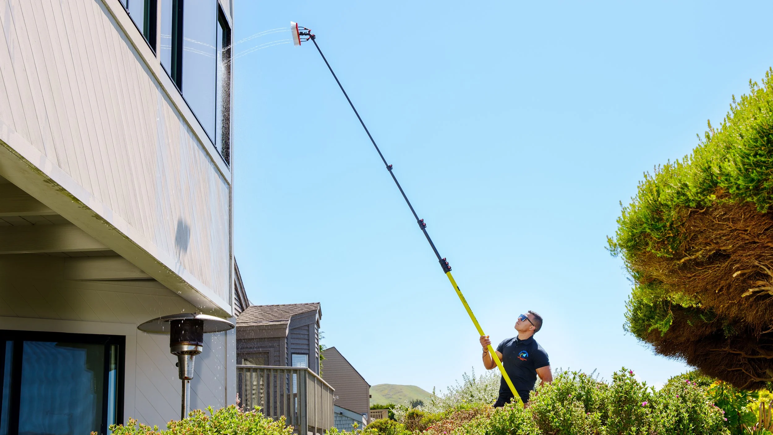 A man using a long extendable tool to clean the exterior windows of a multi-story house on a clear, sunny day.