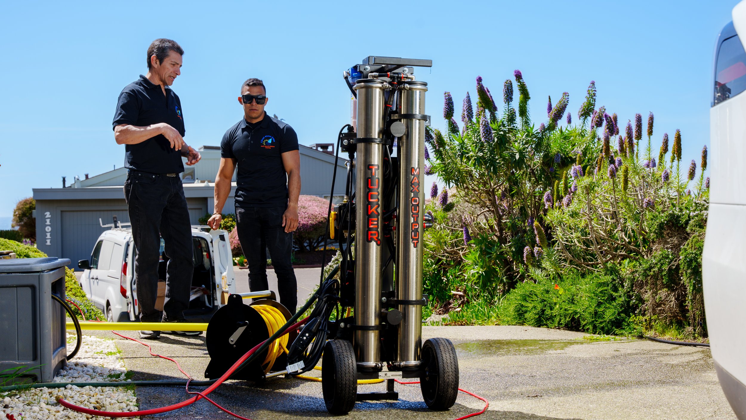 Two men operate a large machine outdoors near some plants and a driveway.