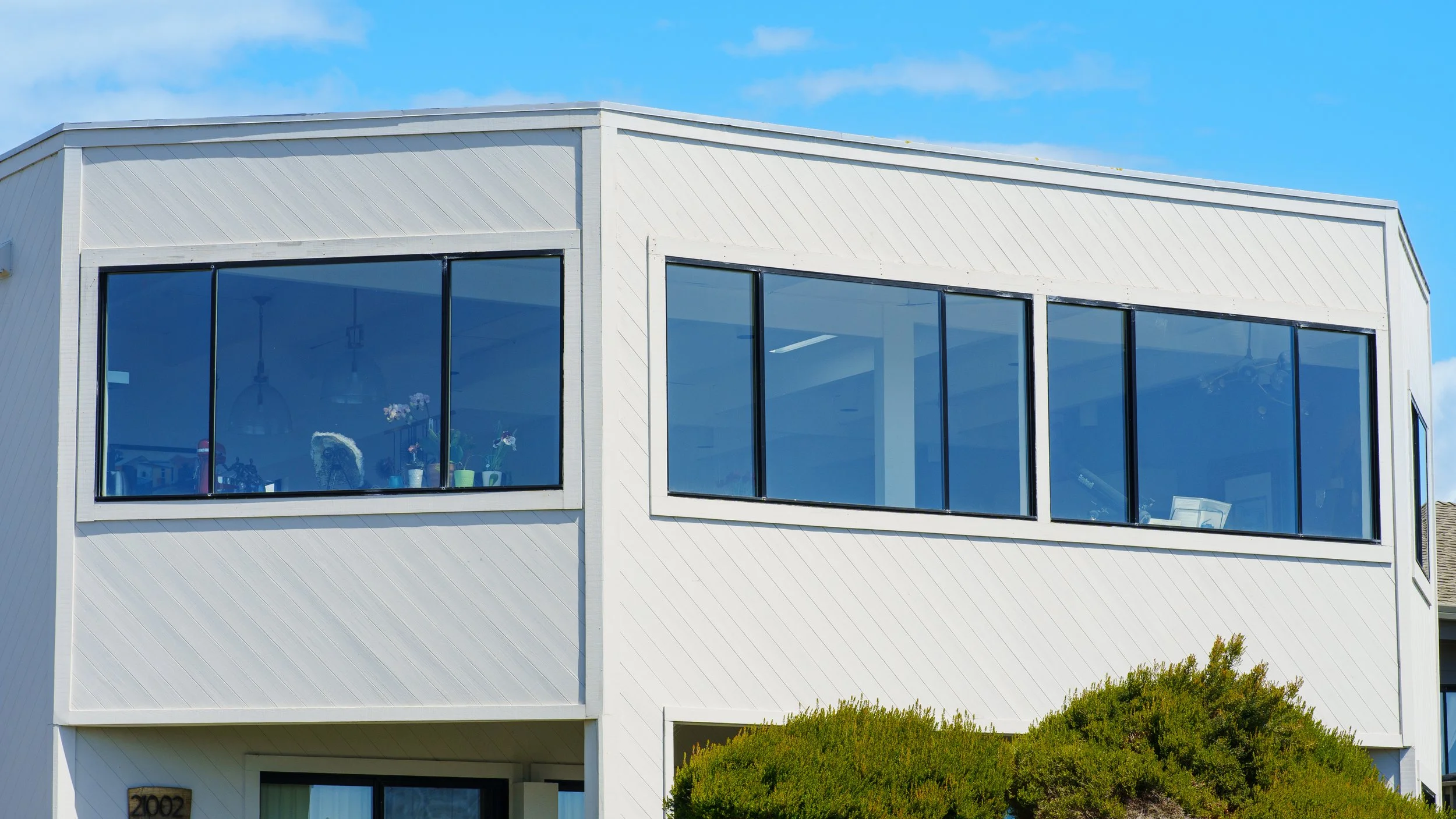 Modern white house with large blue-tinted windows and a partial view of greenery below, under a blue sky.