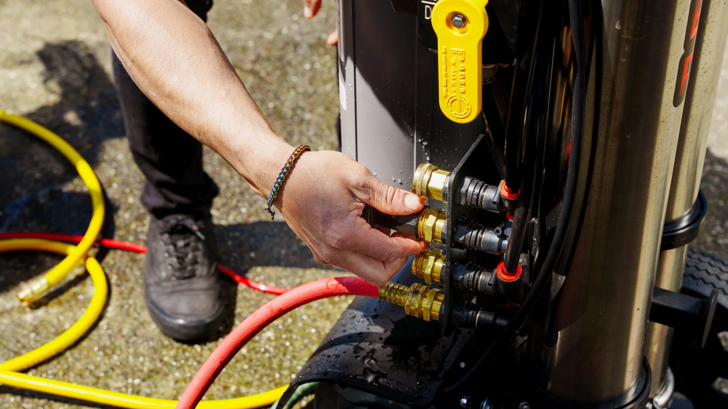 Close-up of a person's hands connecting hoses and fittings to a piece of industrial equipment or machinery outdoors, with yellow, red, and black hoses visible on the ground.