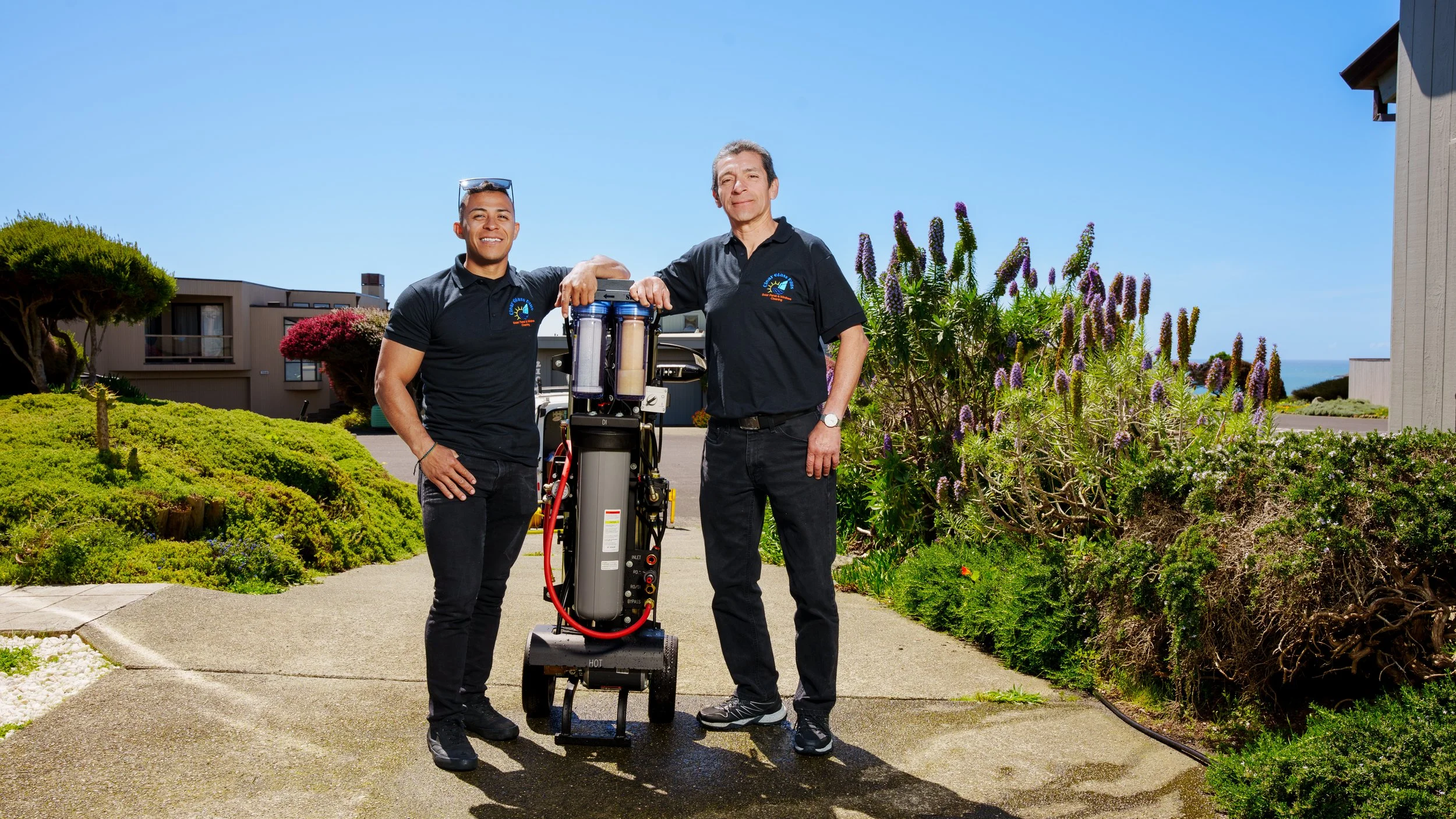 Two men standing outdoors on a sidewalk with plants and shrubs behind them, smiling, one with a water filter system, under clear blue sky.