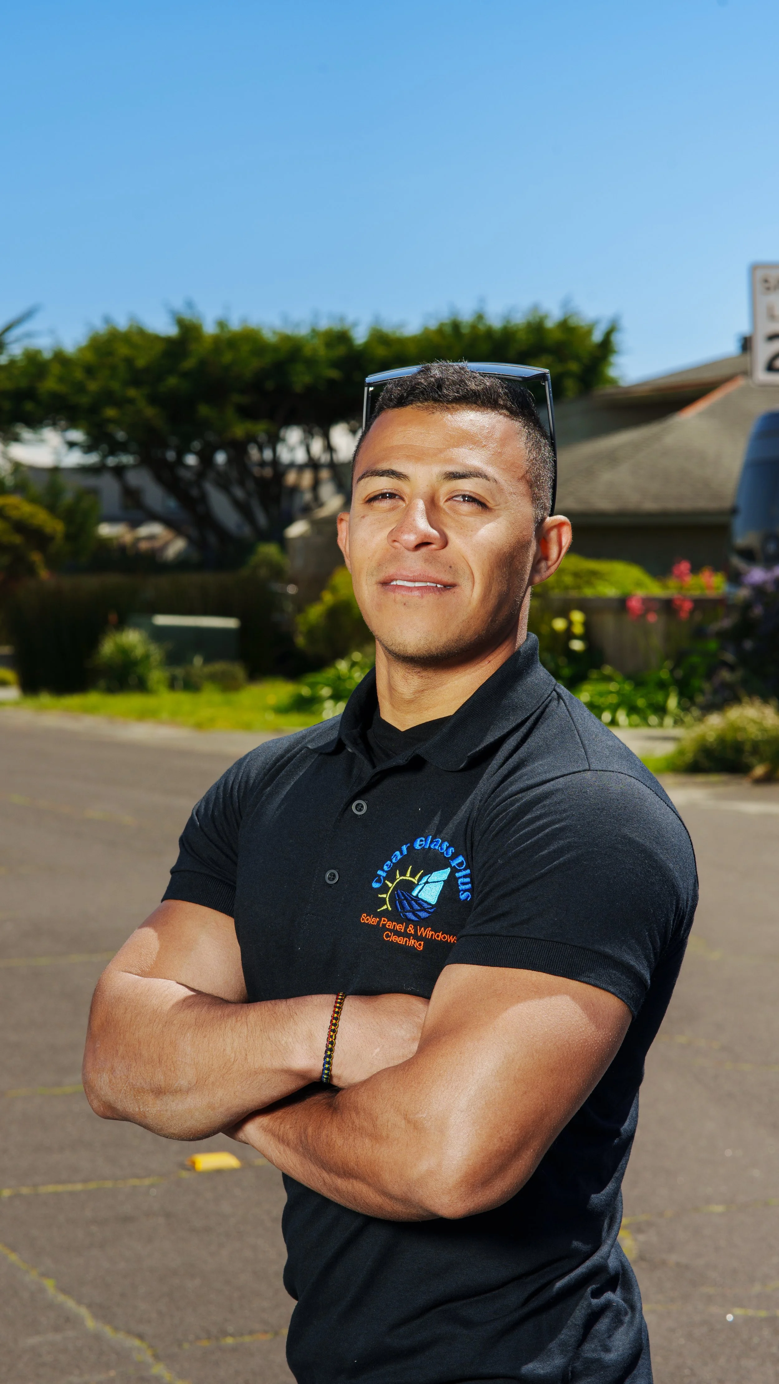 A young man with short dark hair, wearing sunglasses on his head, a black polo shirt with a company logo, standing outdoors with his arms crossed. Background includes trees, a house, and a parking lot.