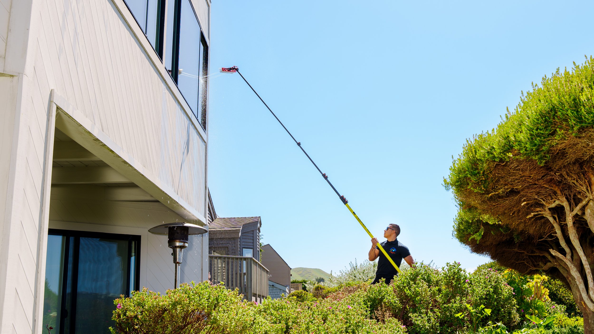 A man is using a long pole to clean or reach the window of a white house, with a clear blue sky and green trees in the background.