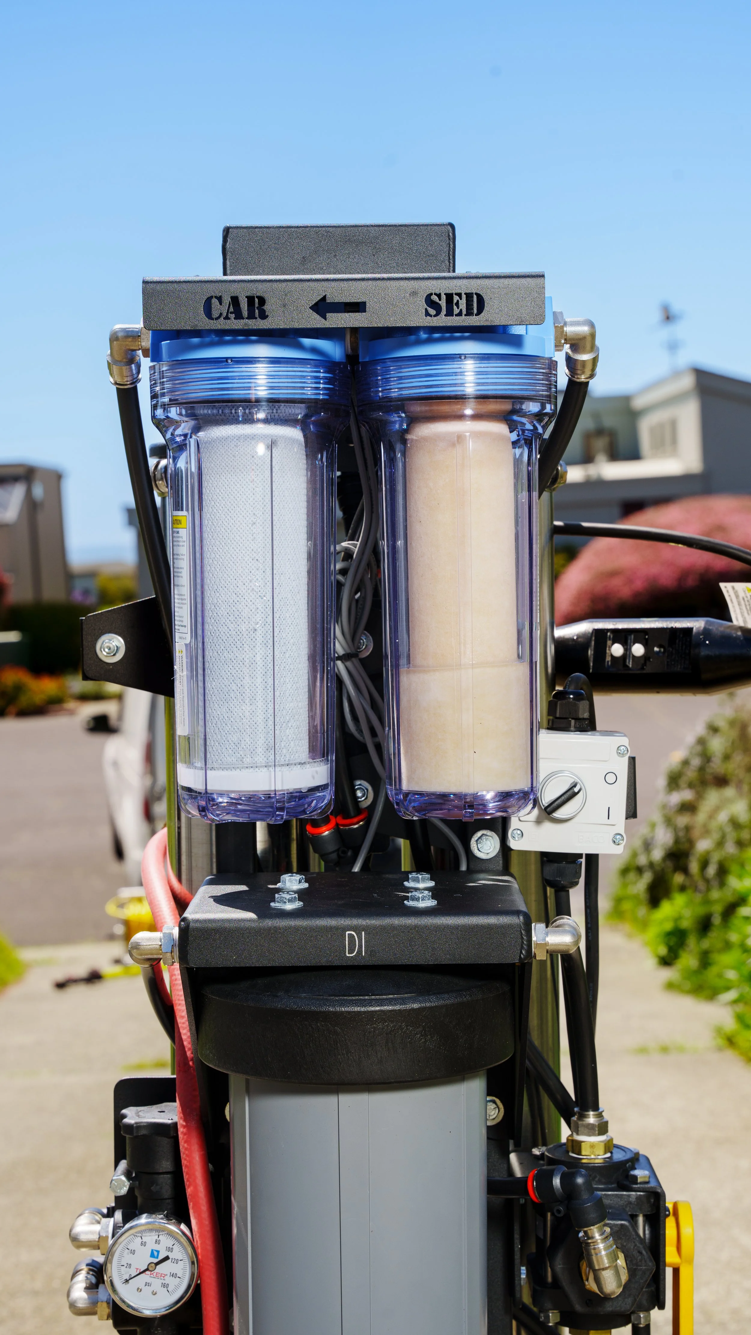 A filtration system with two clear vertical filter housings, one containing a white filter and the other a tan filter, mounted on a metal frame with labels indicating 'CAR' and 'SED' with an arrow pointing left. The system features various tubes, a p