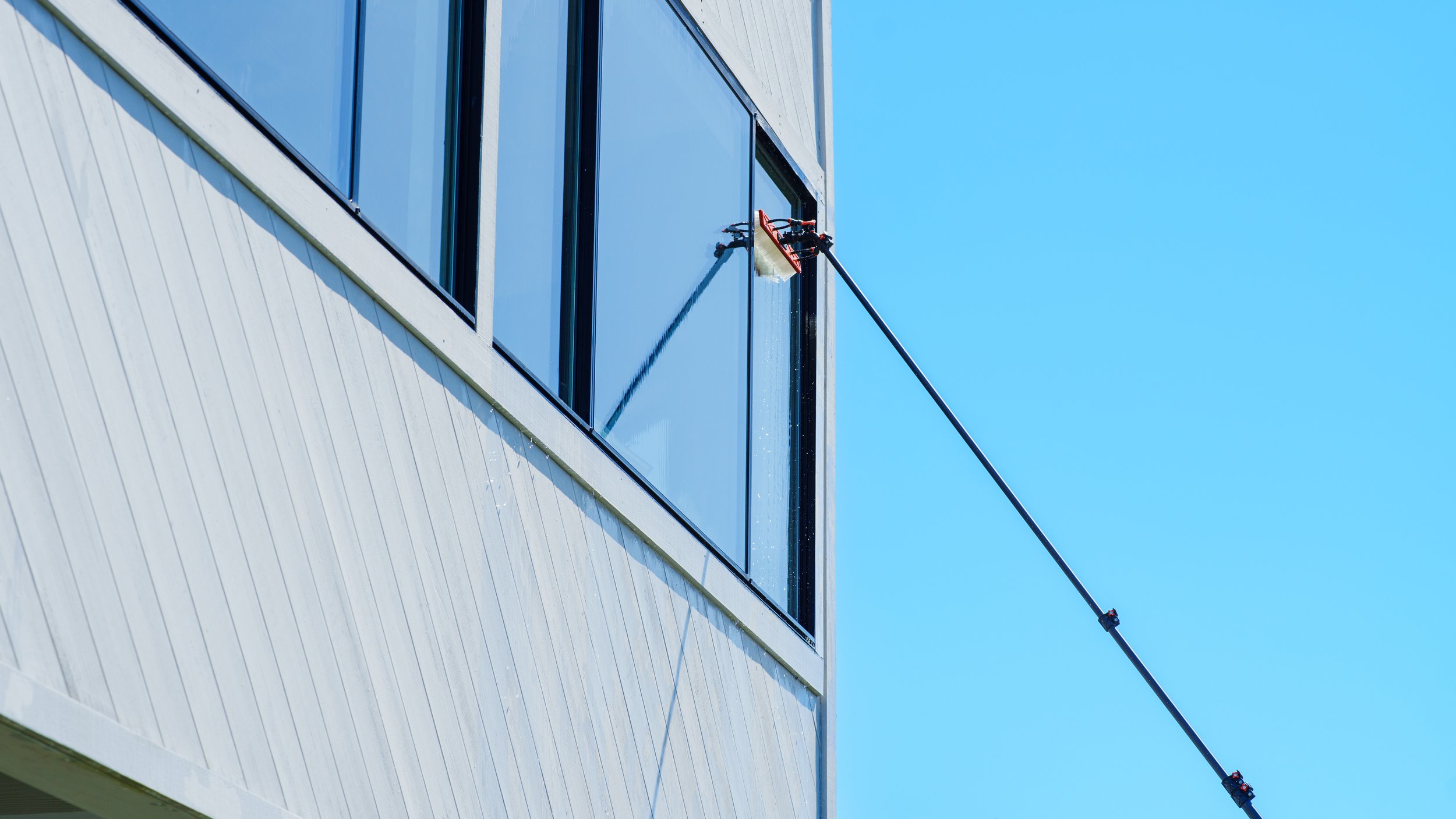 Window cleaning tool attached to a long pole cleaning the exterior glass of a modern building against a clear blue sky.