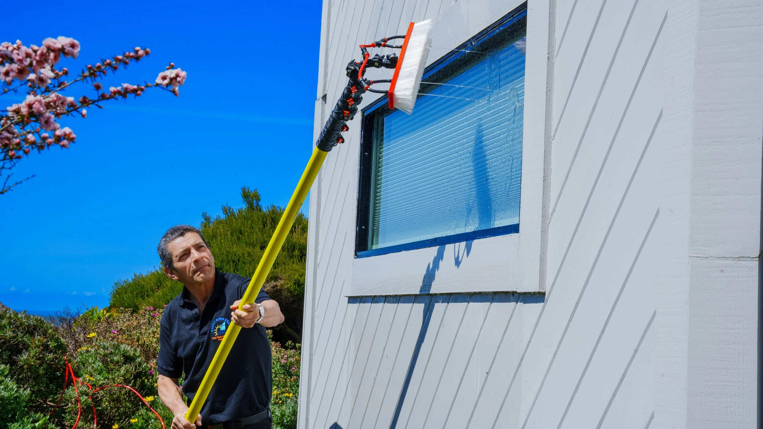 Man cleaning lower window of a white house with a long yellow pole in a sunny garden with blooming trees and bushes.