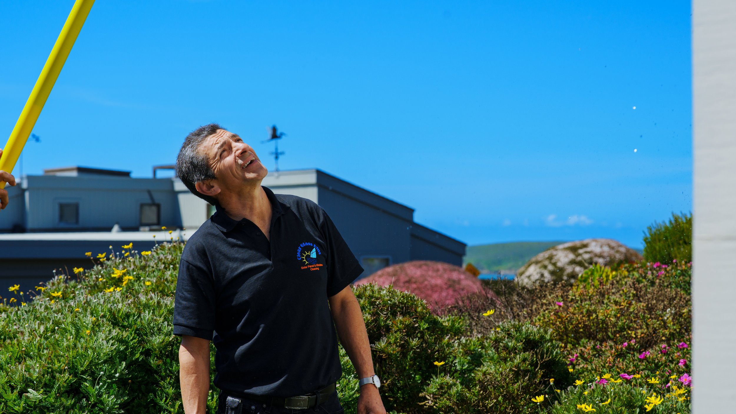 A man smiling outdoors in a sunny garden, surrounded by colorful flowers and bushes, with a building and a clear blue sky in the background.
