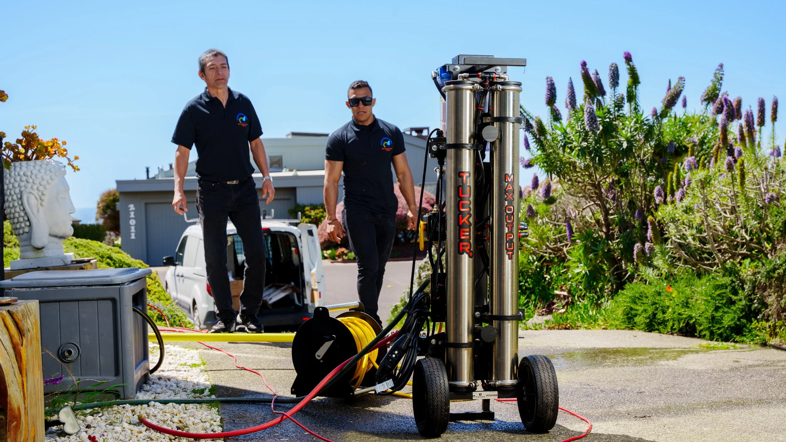Two workers in black shirts working outdoors with a large machine, plants, and a parked white vehicle nearby on a sunny day.