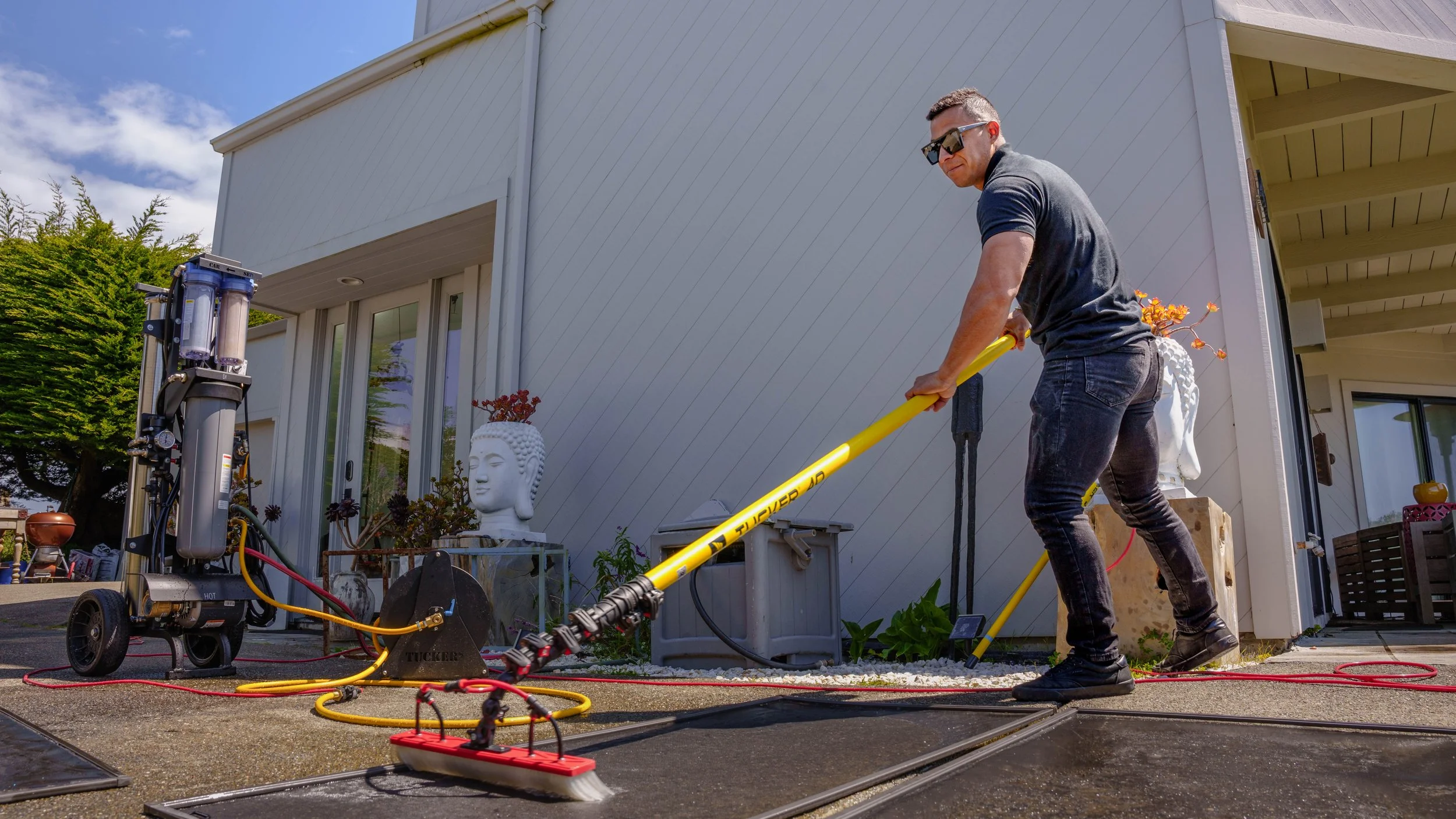 A man wearing sunglasses and a dark t-shirt is using a soil tiller in a backyard. There are gardening statues and a piece of equipment with filters nearby, and the house has white siding and large windows.