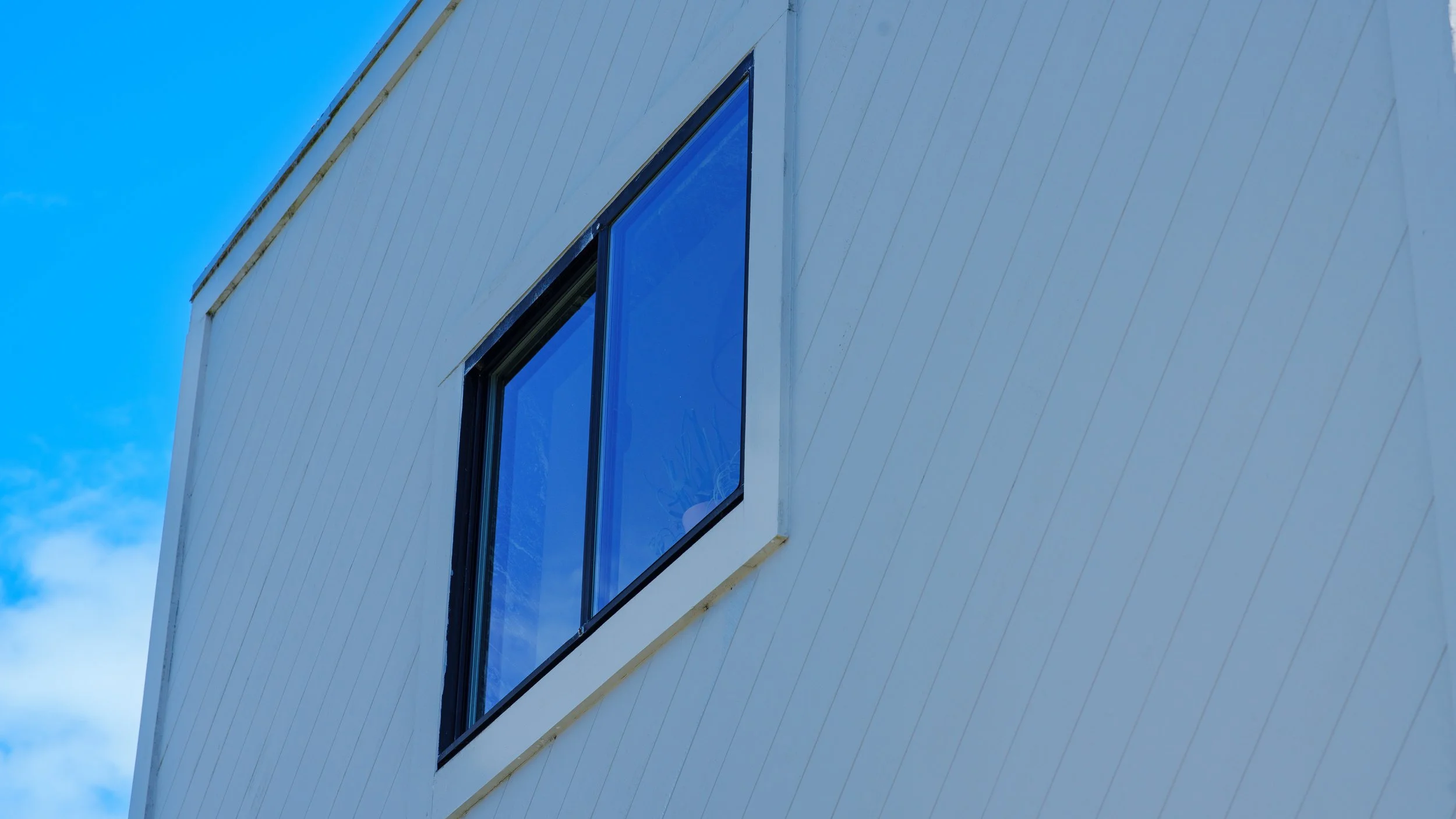 Close-up view of a modern building's exterior corner with white siding and two rectangular, dark-framed windows reflecting the blue sky.