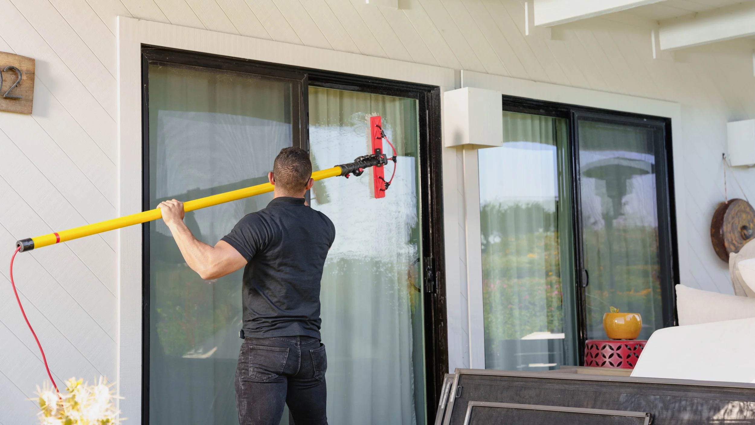 A man is cleaning large glass sliding doors outside with a long-handled squeegee.