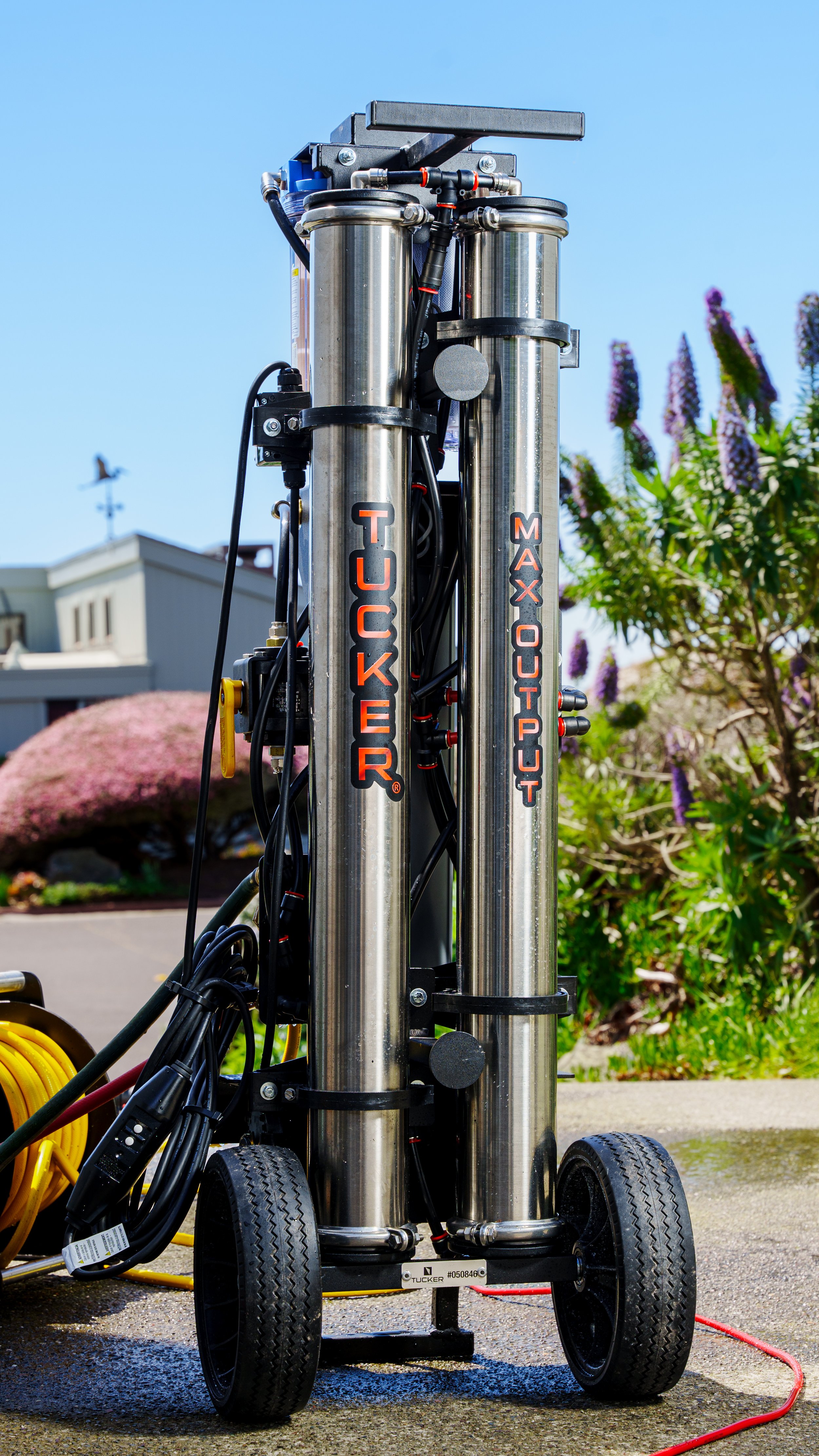 A metal water jetting cleaning machine labeled 'Tucker Max Out' with black wheels and connected hoses, outdoors on a paved surface with greenery and buildings in the background.