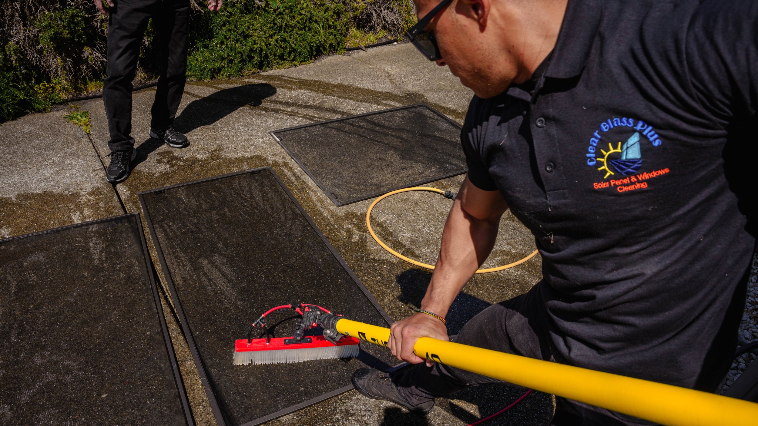 Two workers cleaning solar panels outdoors with cleaning tools, one wearing a black shirt with a company logo.