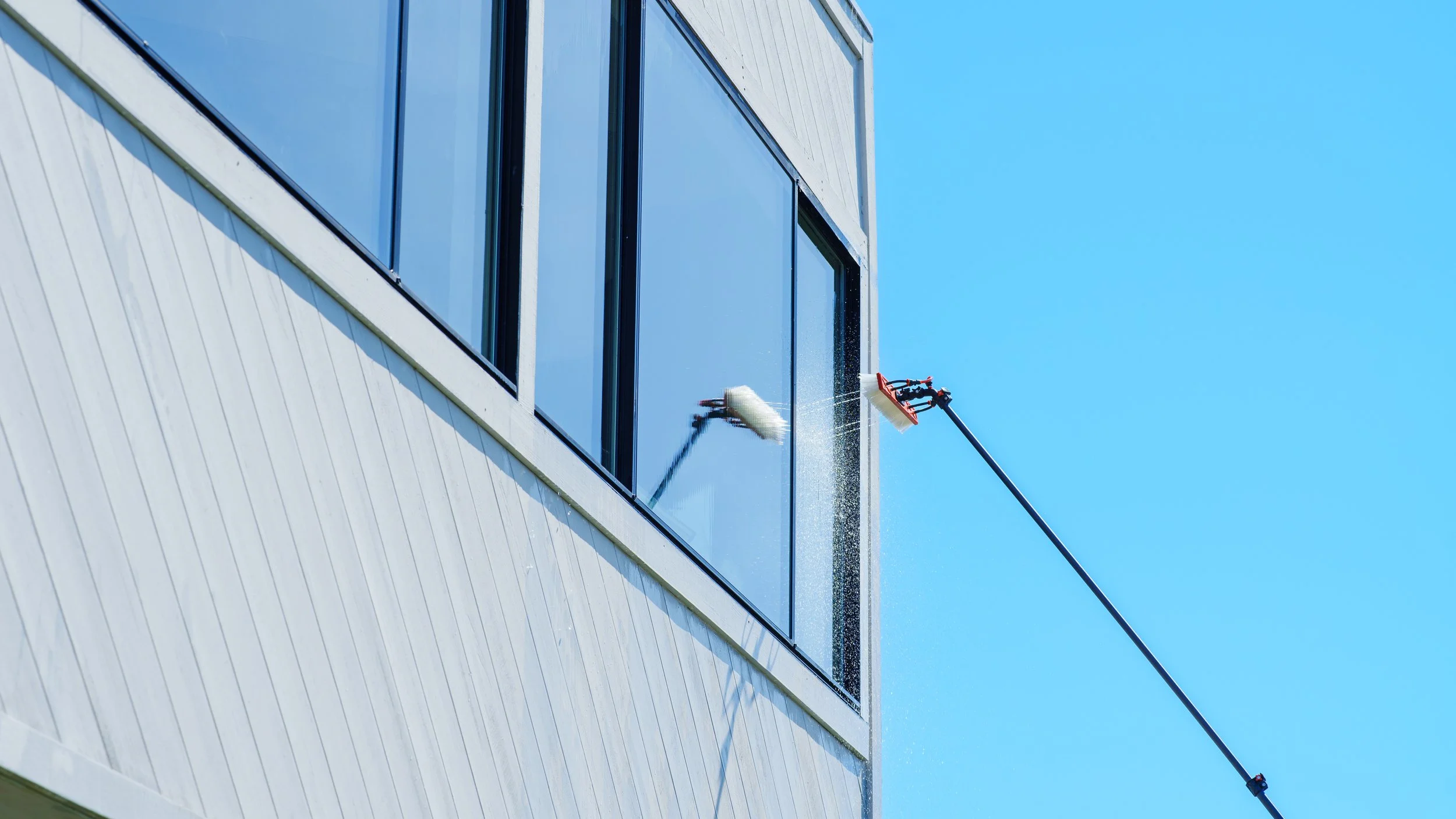 A person cleaning a large window on the upper floor of a modern building with a long pole and roller brush, against a clear blue sky.