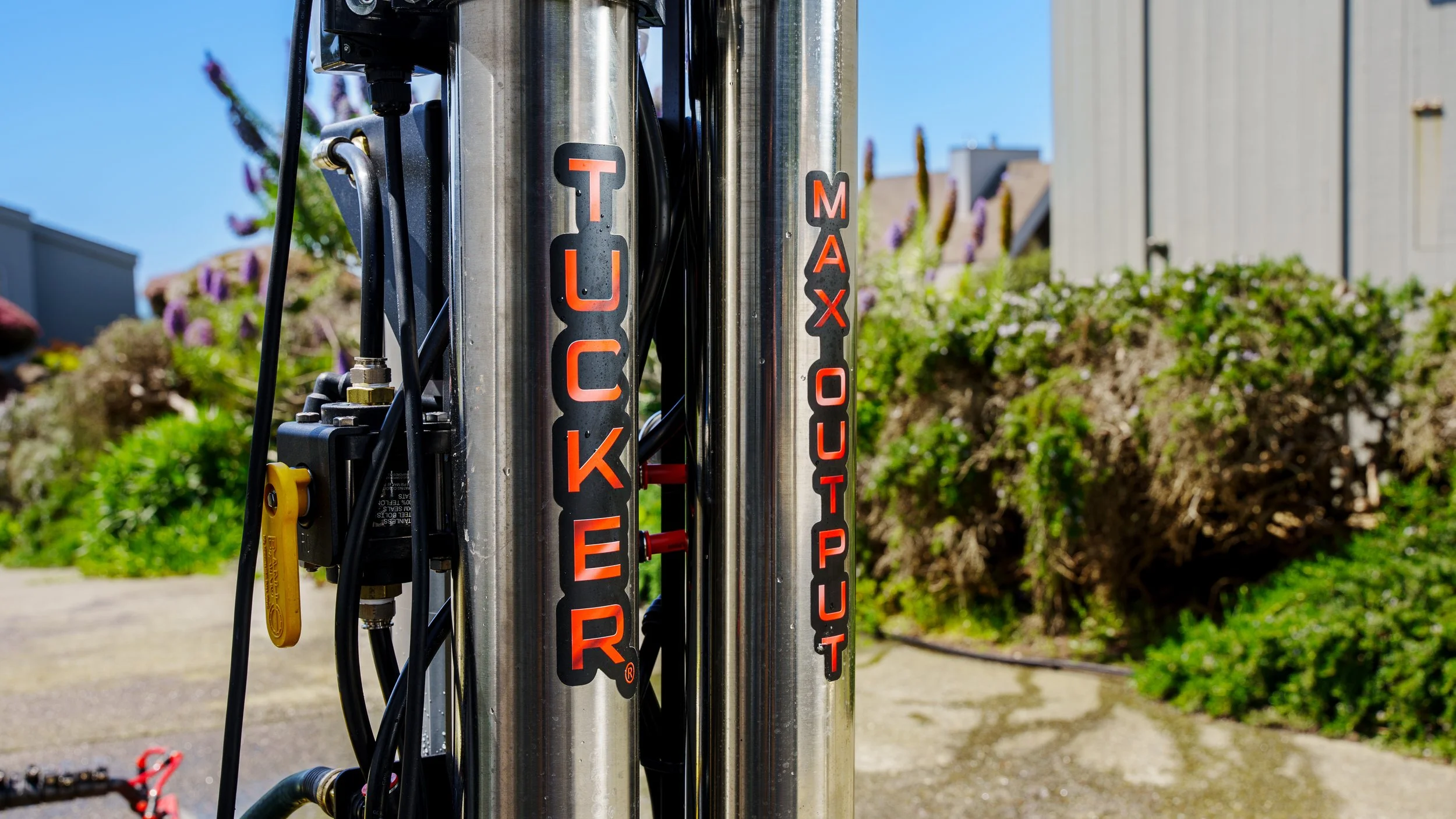 Close-up of a vertical metal structure with black and red stickers reading 'TOUCH' and 'MAX' and various cables attached, outdoors with a background of buildings and bushes under a clear blue sky.