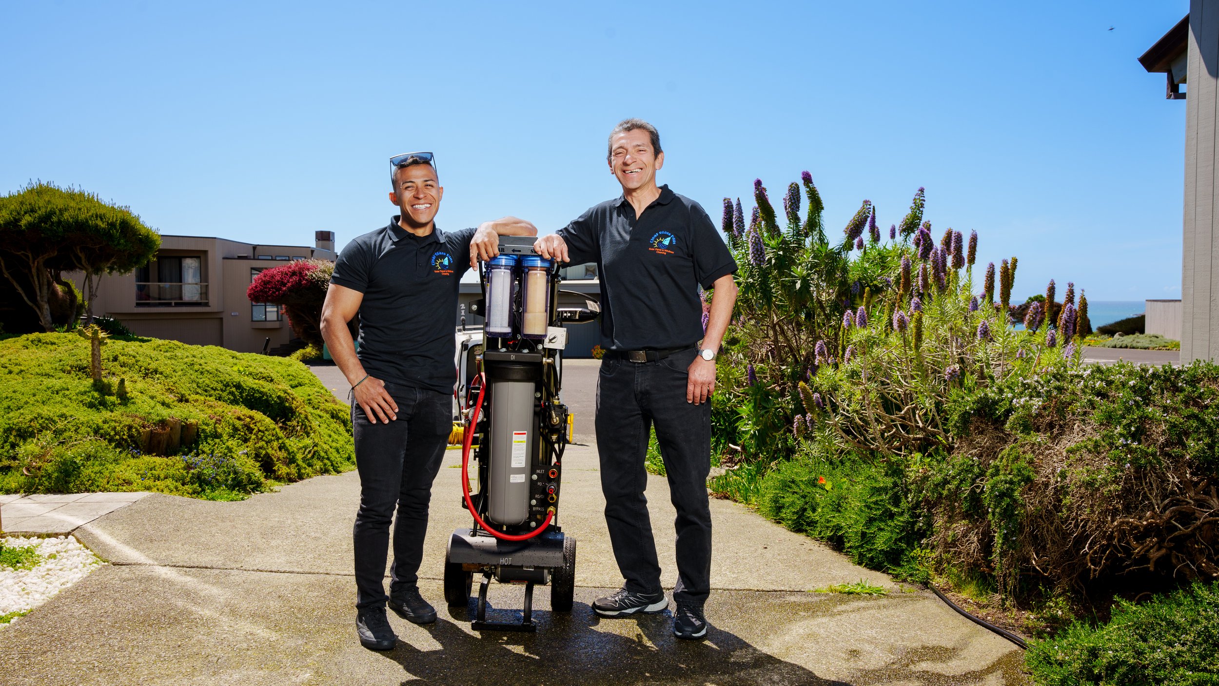 Two men in black polo shirts standing outdoors beside a water filtration system, smiling, with plants and residential buildings in the background on a sunny day.
