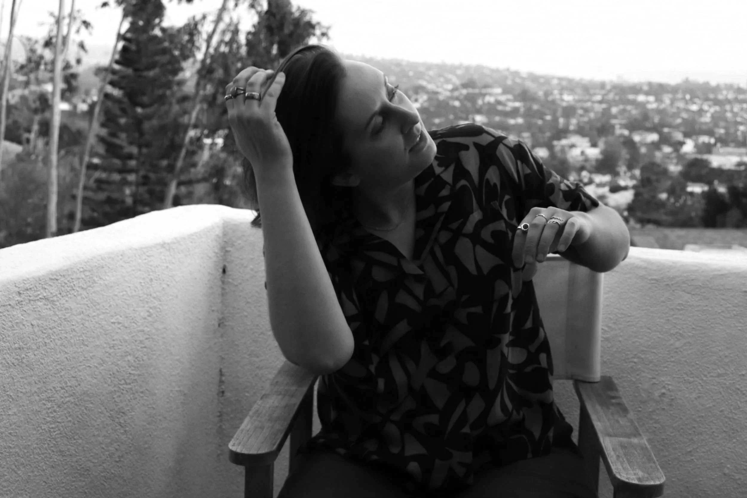 A woman with dark hair and multiple rings on her fingers is sitting at a table outdoors, leaning her head on her hand with eyes closed and a slight smile. The background shows trees and a cityscape in black and white.