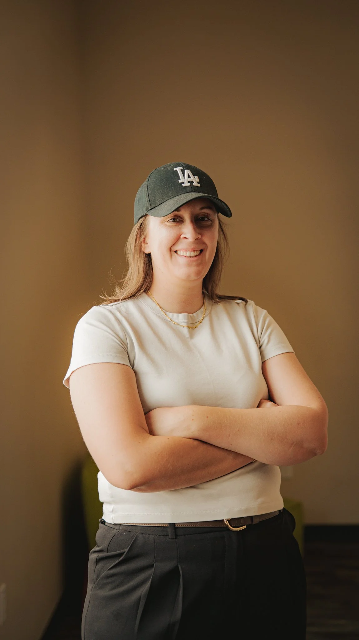 A woman with light brown hair wearing a dark baseball cap with 'LA' logo, a beige t-shirt, and dark pants, standing with arms crossed and smiling against a plain brown background.