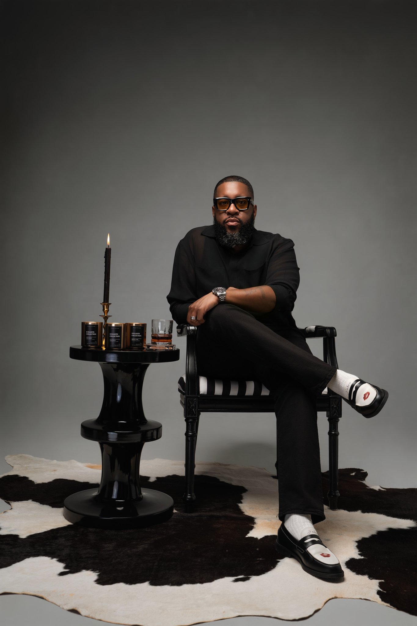 A man with a beard and sunglasses sitting on a black chair, wearing a black shirt, black pants, and white shoes with lips design, in a studio with a gray background. There is a black side table with candles and a glass of whiskey beside him, and a black and white cowhide rug on the floor.